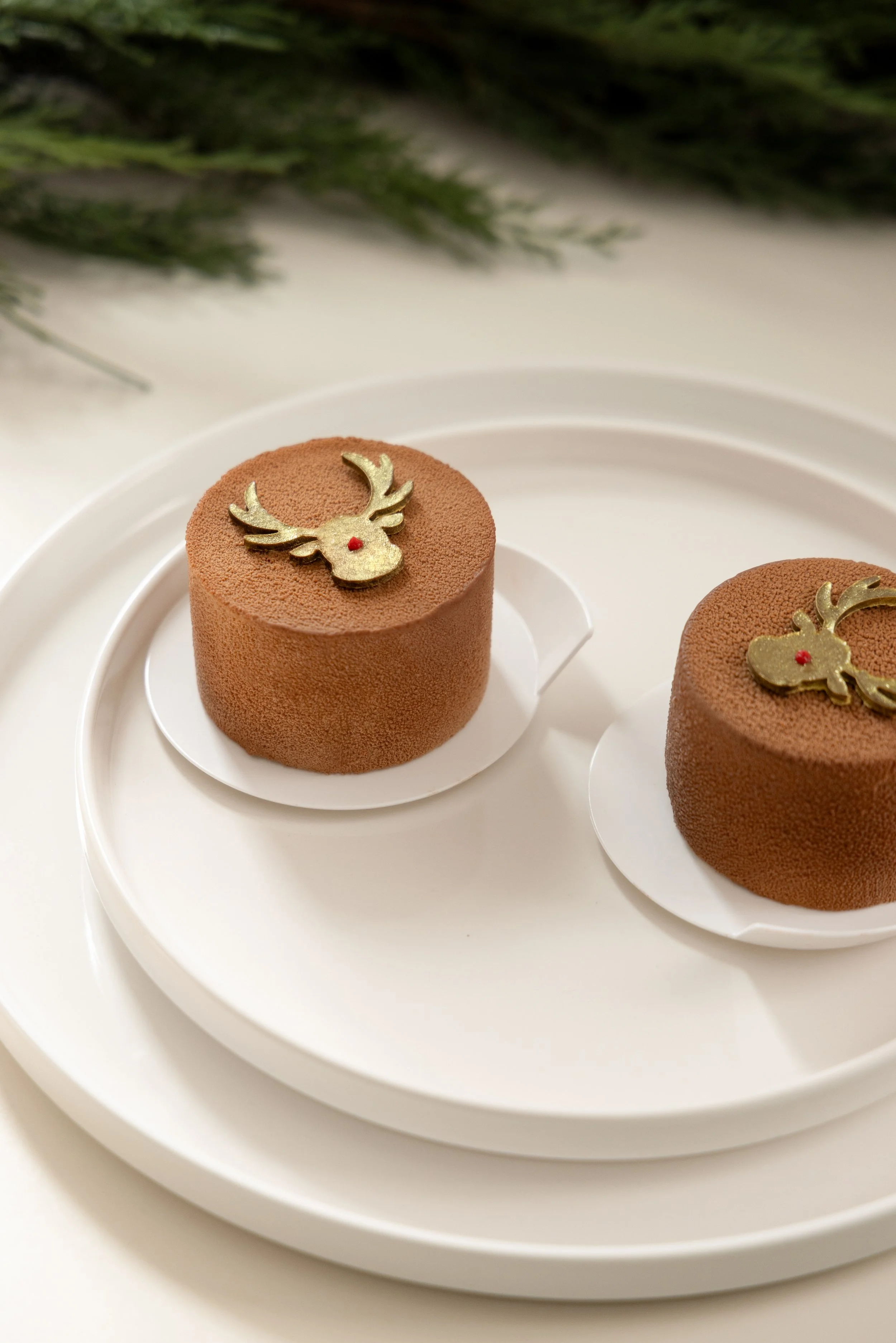 Two small round chocolate desserts with reindeer-shaped gold decorations on top, placed on white circular trays