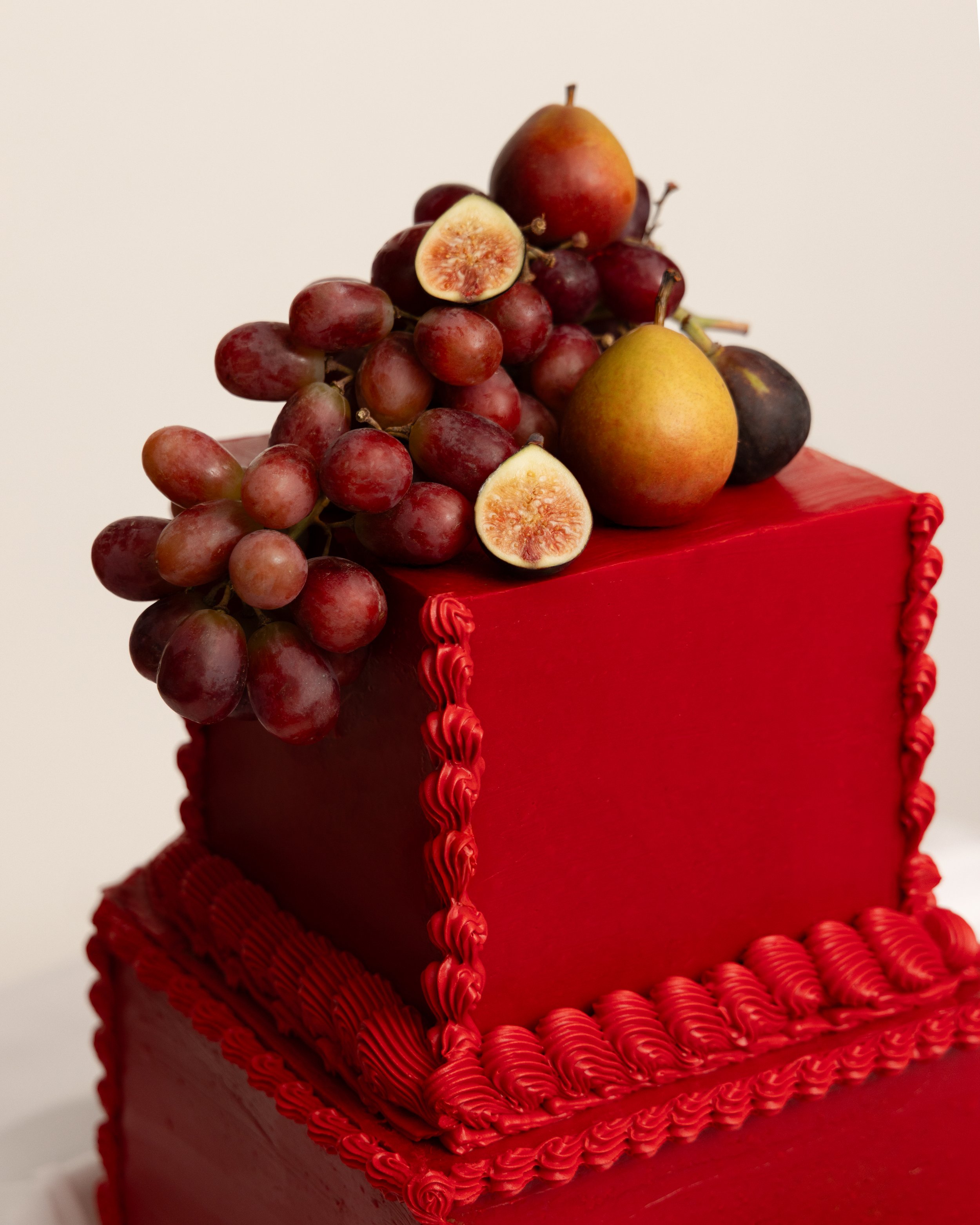 A red cake decorated with red icing and topped with grapes, figs, and pears.