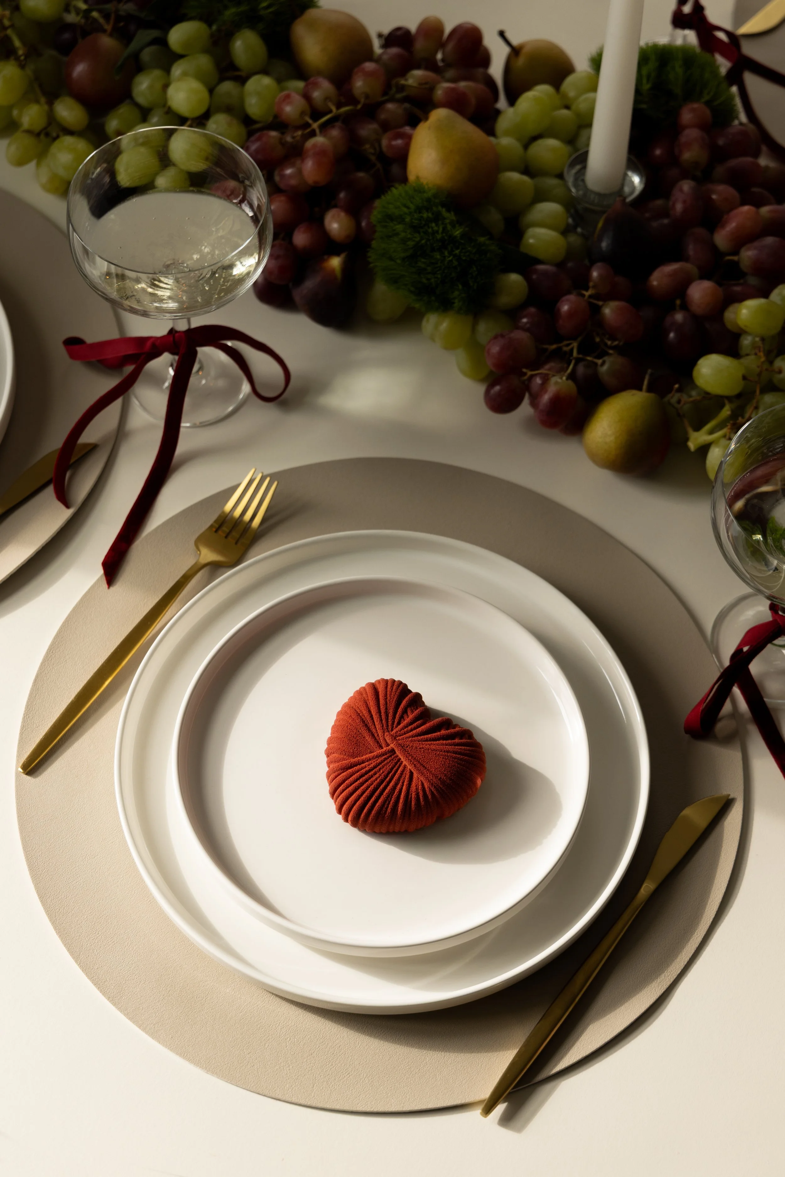 Place setting with white plates, gold fork and knife, glass of water with red ribbon, decorative centerpiece with grapes, pears, apple, moss, and candle.