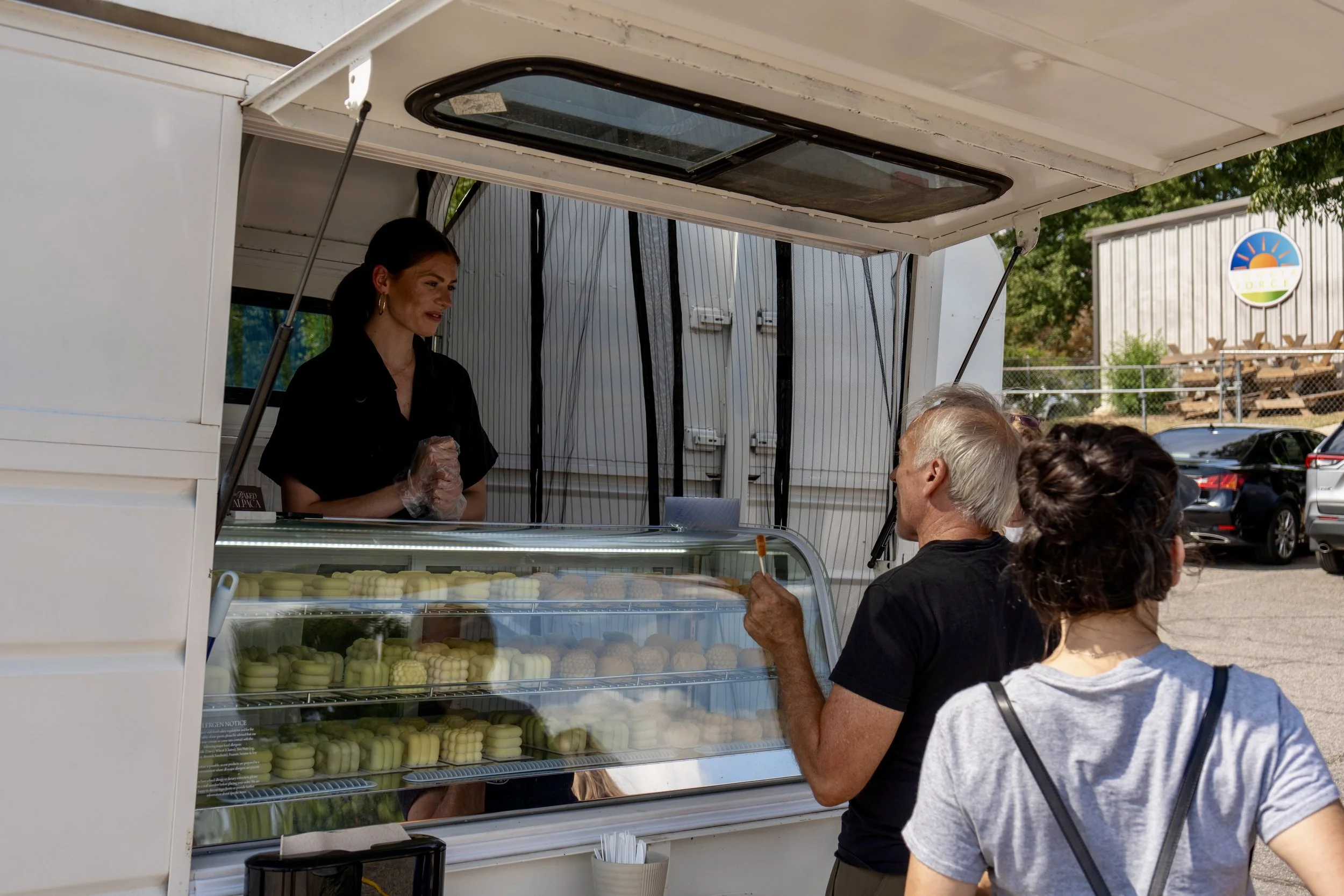 A woman serving ice cream at a mobile ice cream stand to an elderly man and a woman in line, with a parking lot and a building with a sign featuring a sun and rainbow in the background.