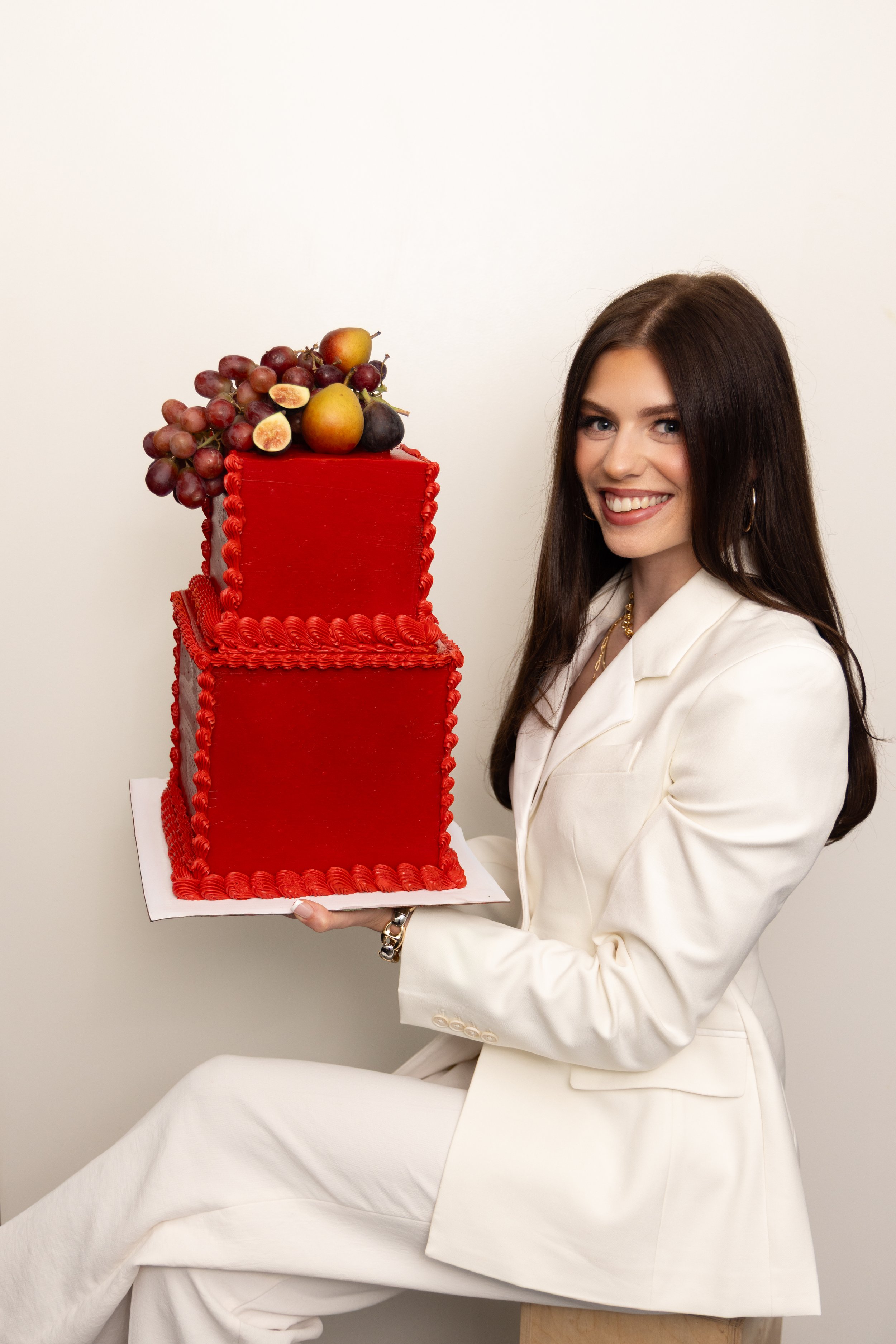 A woman with long dark hair wearing a white suit holds a large red frosted cake decorated with fresh grapes and pears.