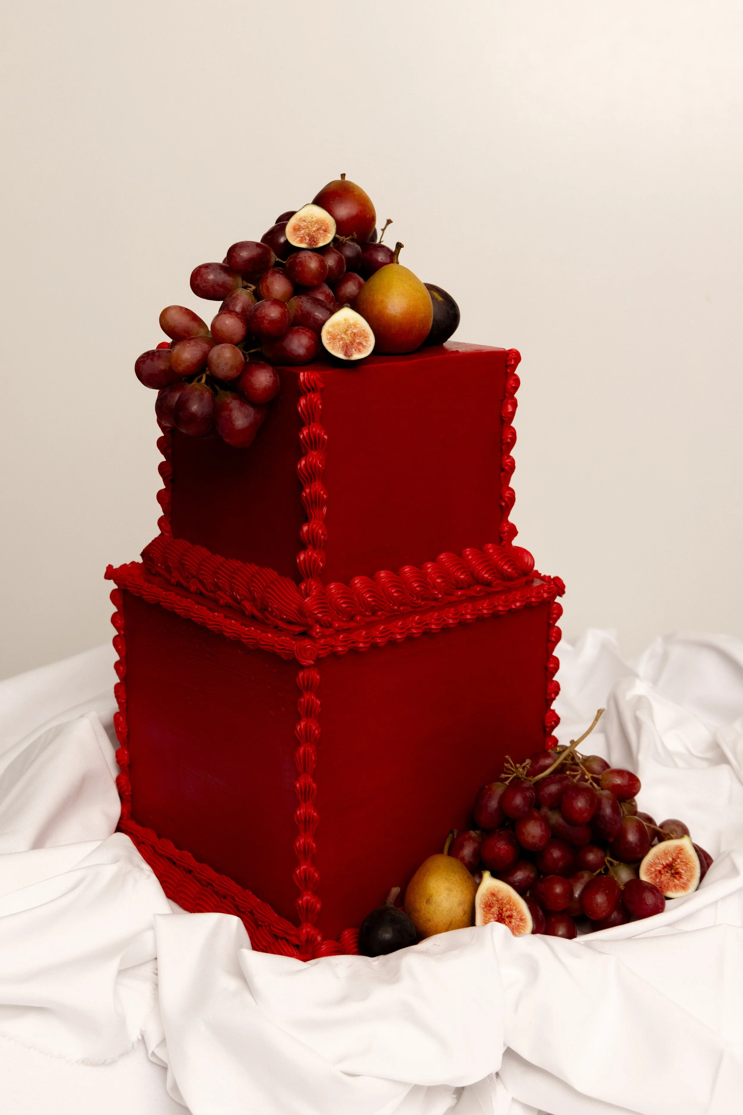 A multi-tiered red cake decorated with red icing, topped with grapes, figs, pears, and plums, with additional fruits around the base, all set on white fabric against a plain background.