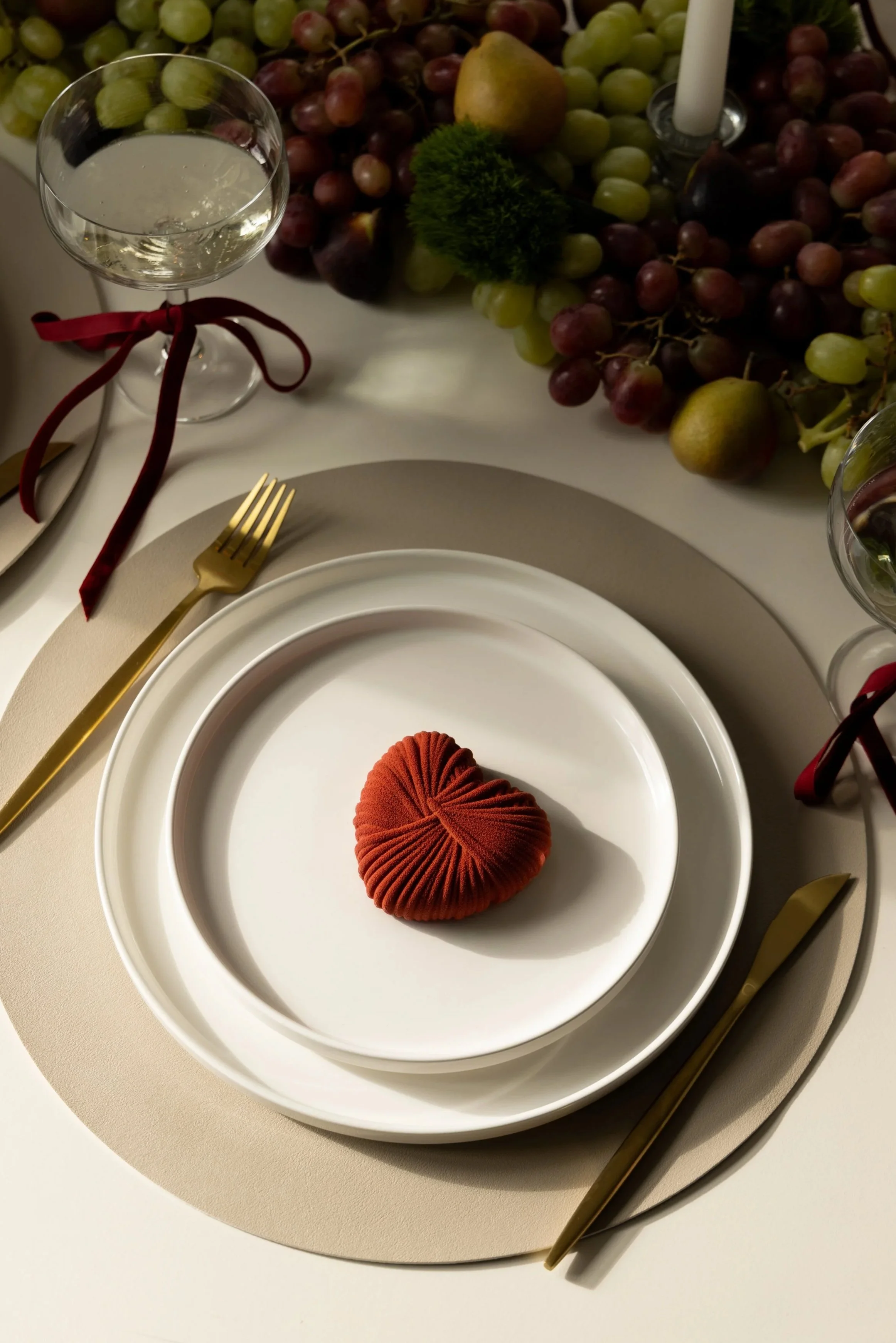 Elegant table setting with white plates, a gold fork and knife, a wine glass with white wine, and a decorative red cloth napkin, with a centerpiece of grapes, fruit, greenery, and a candle.