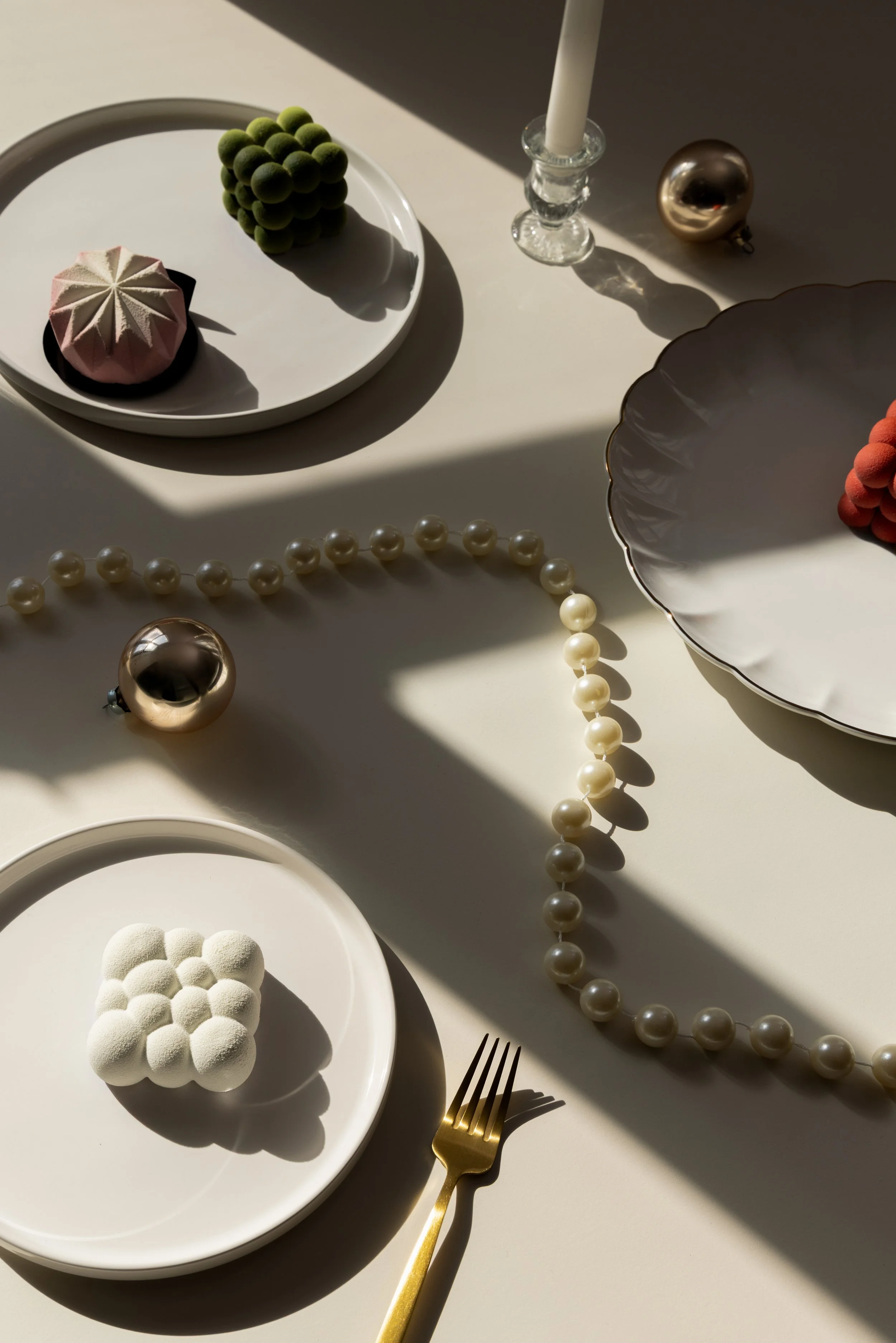 Table setting with decorative plates, a pearl necklace, a gold fork, silver and gold ornaments, a white candle in a glass holder, and assorted colorful desserts, with sunlight casting shadows.