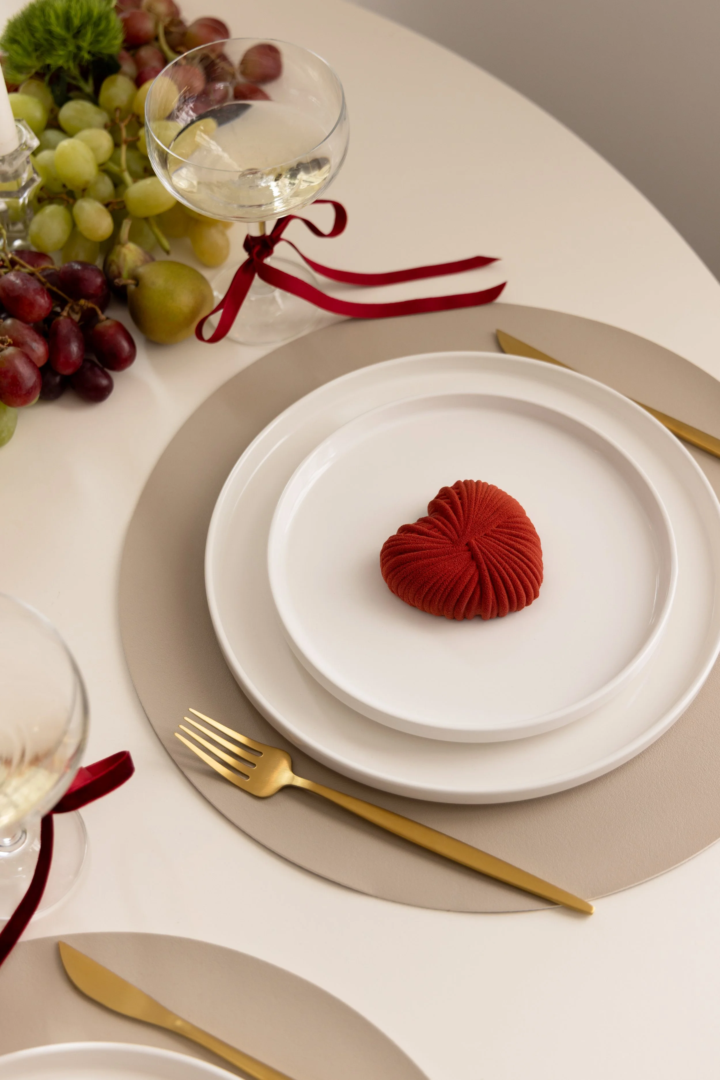 A table setting with a white plate, a smaller white plate, and a red cloth-shaped decoration on top. Gold-colored fork and knife are placed beside the plates. A glass of water with a red ribbon tied around its stem is also on the table. There are clusters of green and purple grapes and a small glass container with a cork lid in the background.