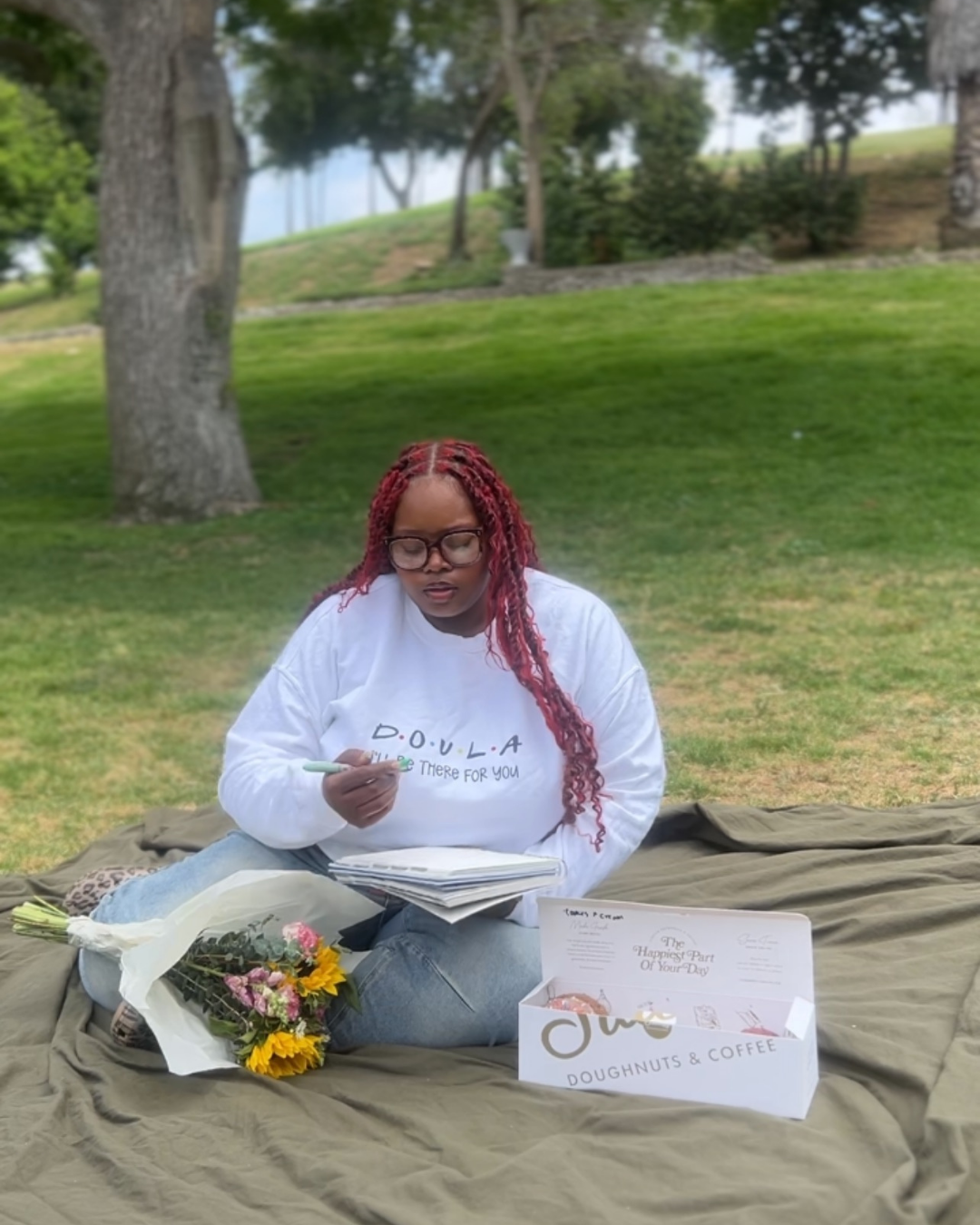 A woman with red braided hair and glasses sitting on a blanket outdoors, holding a pen and a notebook, with flowers and a box of donuts and coffee nearby.