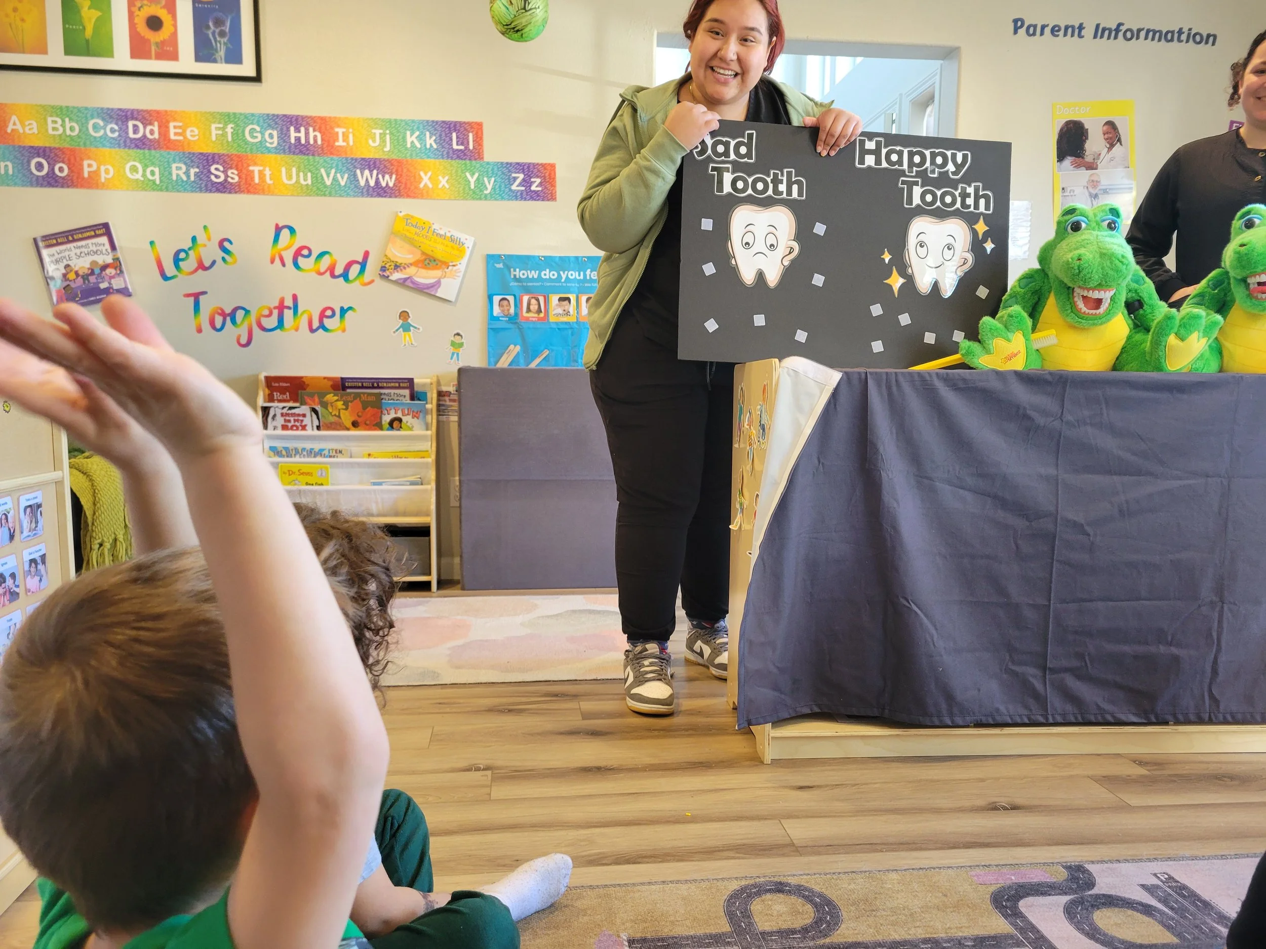 A teacher holding a sign with a tooth illustration comparing a sad tooth and a happy tooth in a classroom, with children sitting on the floor raising their hands.