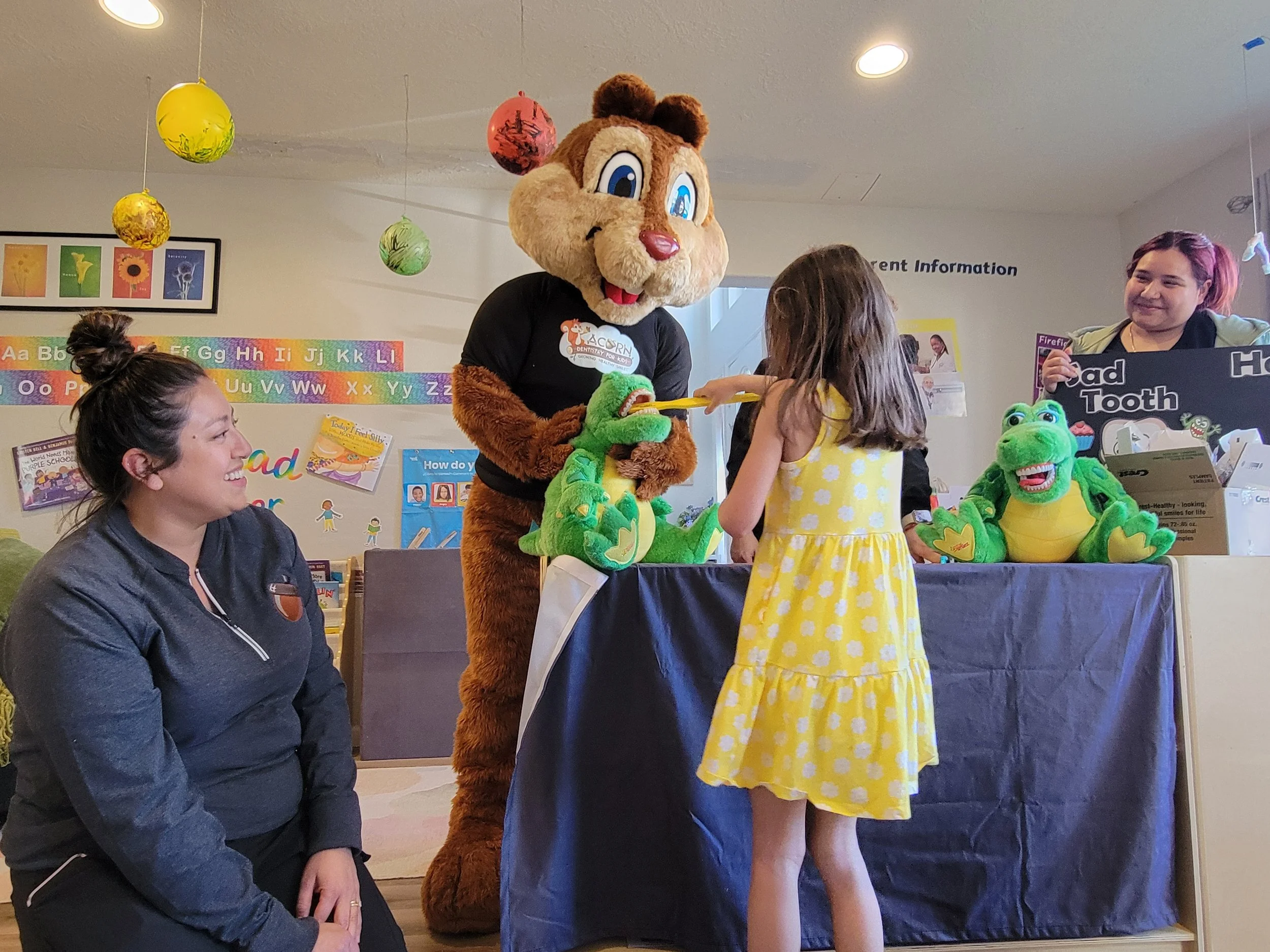 A young girl in a yellow dress receives a medal from a person in a large lion mascot costume behind a table surrounded by plush crocodile toys, with two women nearby, one sitting and smiling and the other standing behind. The scene appears to be in a classroom decorated with colorful paper lanterns and educational posters.