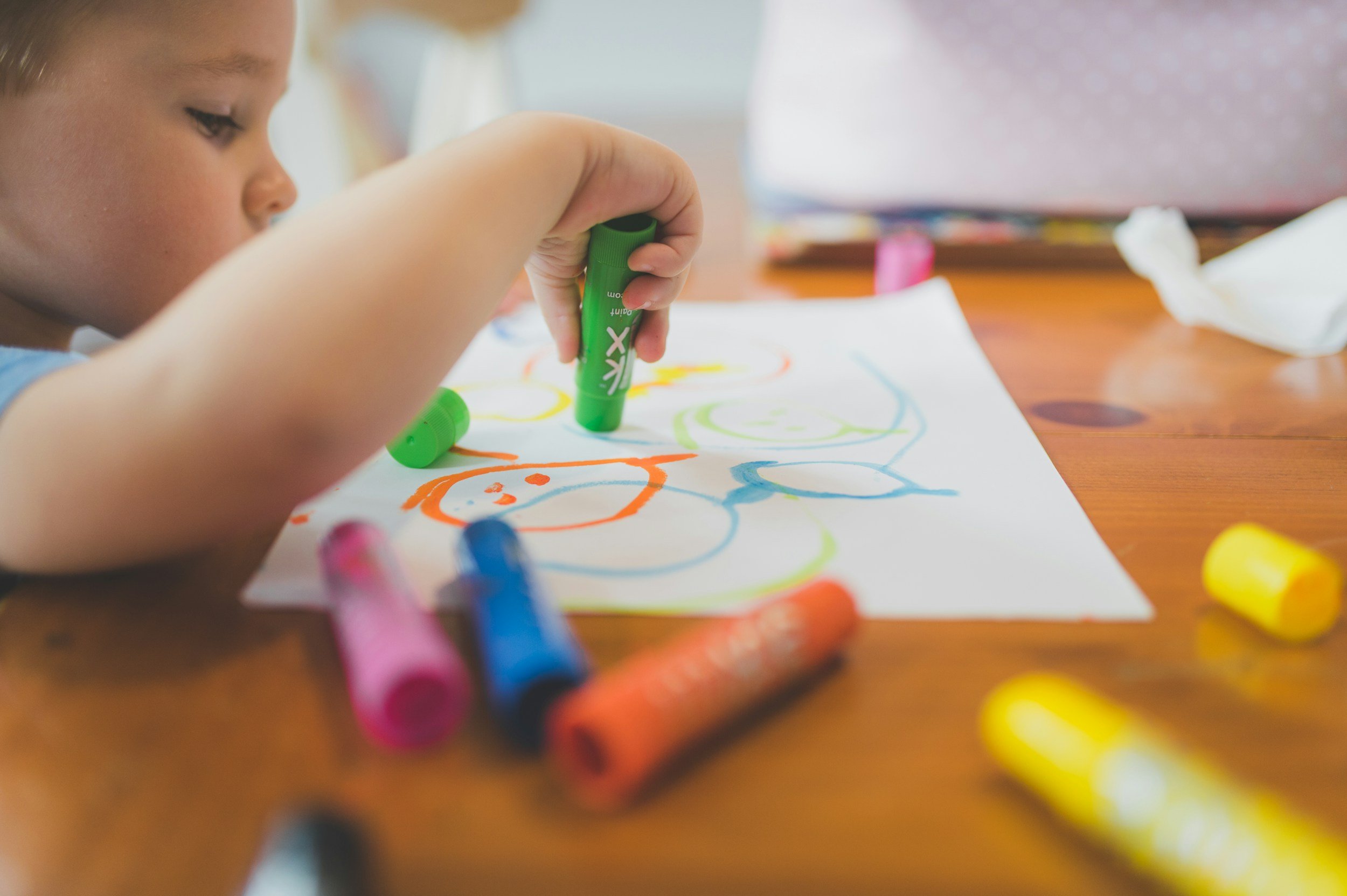 A young child is drawing on a sheet of paper with colorful markers, creating a picture with various shapes and faces. Several markers are scattered on the wooden table.