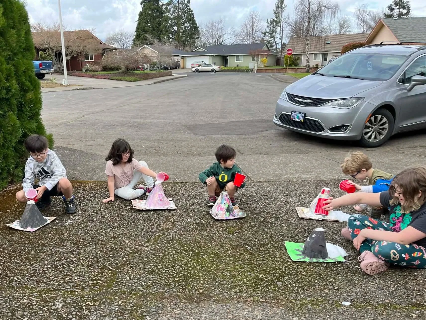 Five children, sitting and kneeling, participate in a science experiment outdoors. They are using red cups and small volcano models on painted bases, with some volcanoes erupting with a fizzy substance. The setting is a residential neighborhood parki