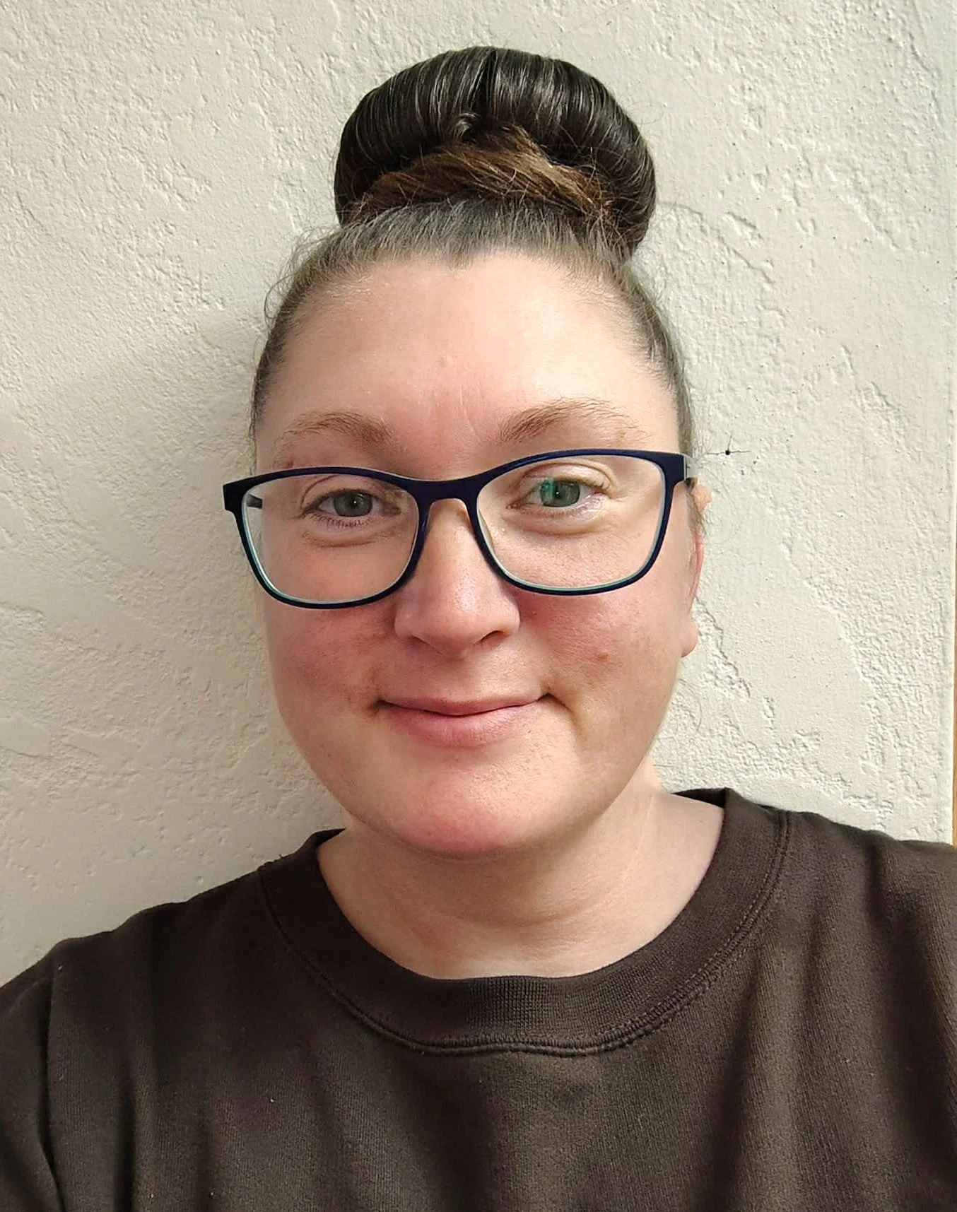 Close-up of a woman with glasses, dark brown hair in a top bun, wearing a dark brown shirt, smiling in front of a textured white wall.