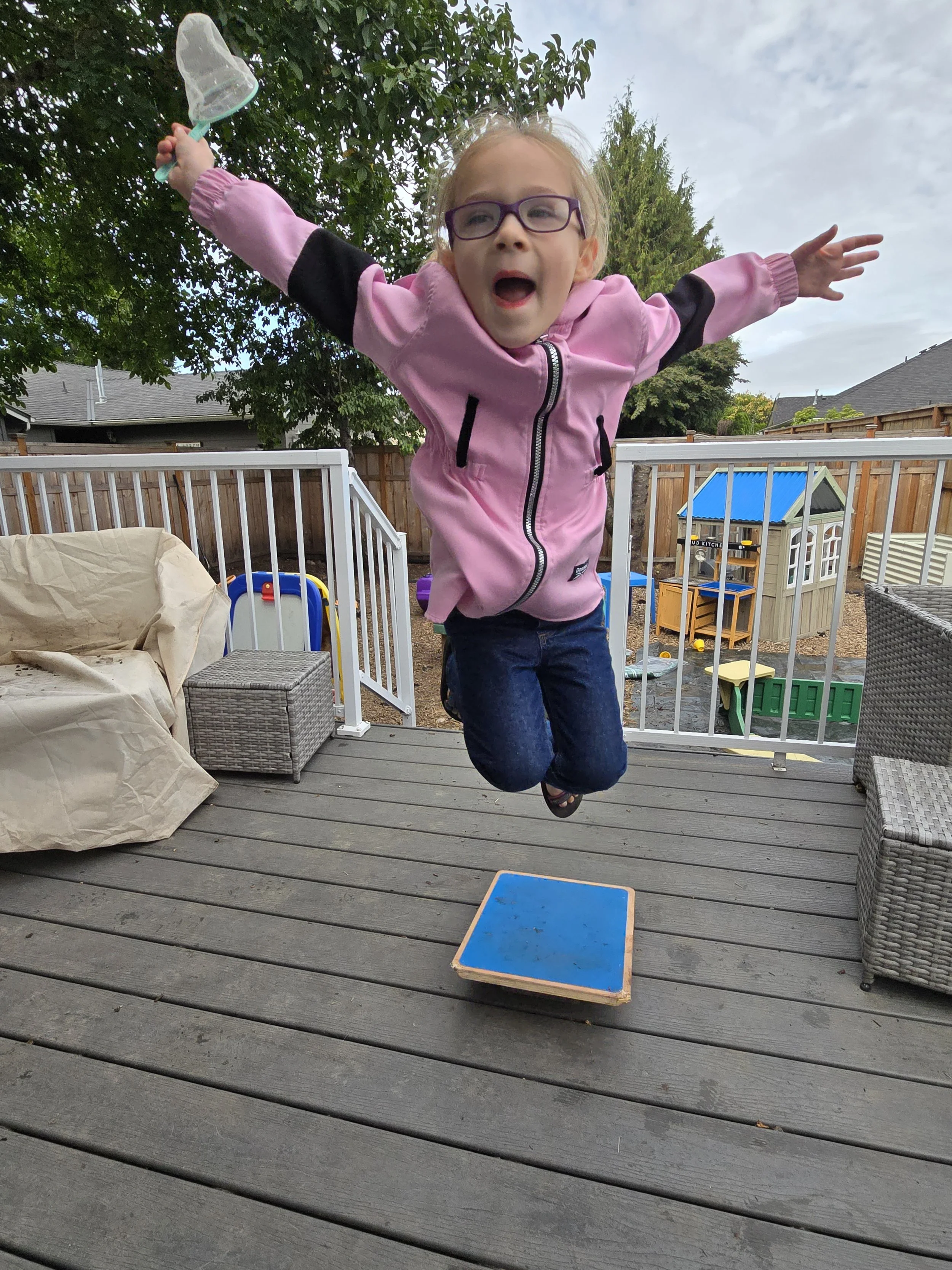 Young girl jumping on a wooden deck, with outdoor furniture and playhouse in the background.