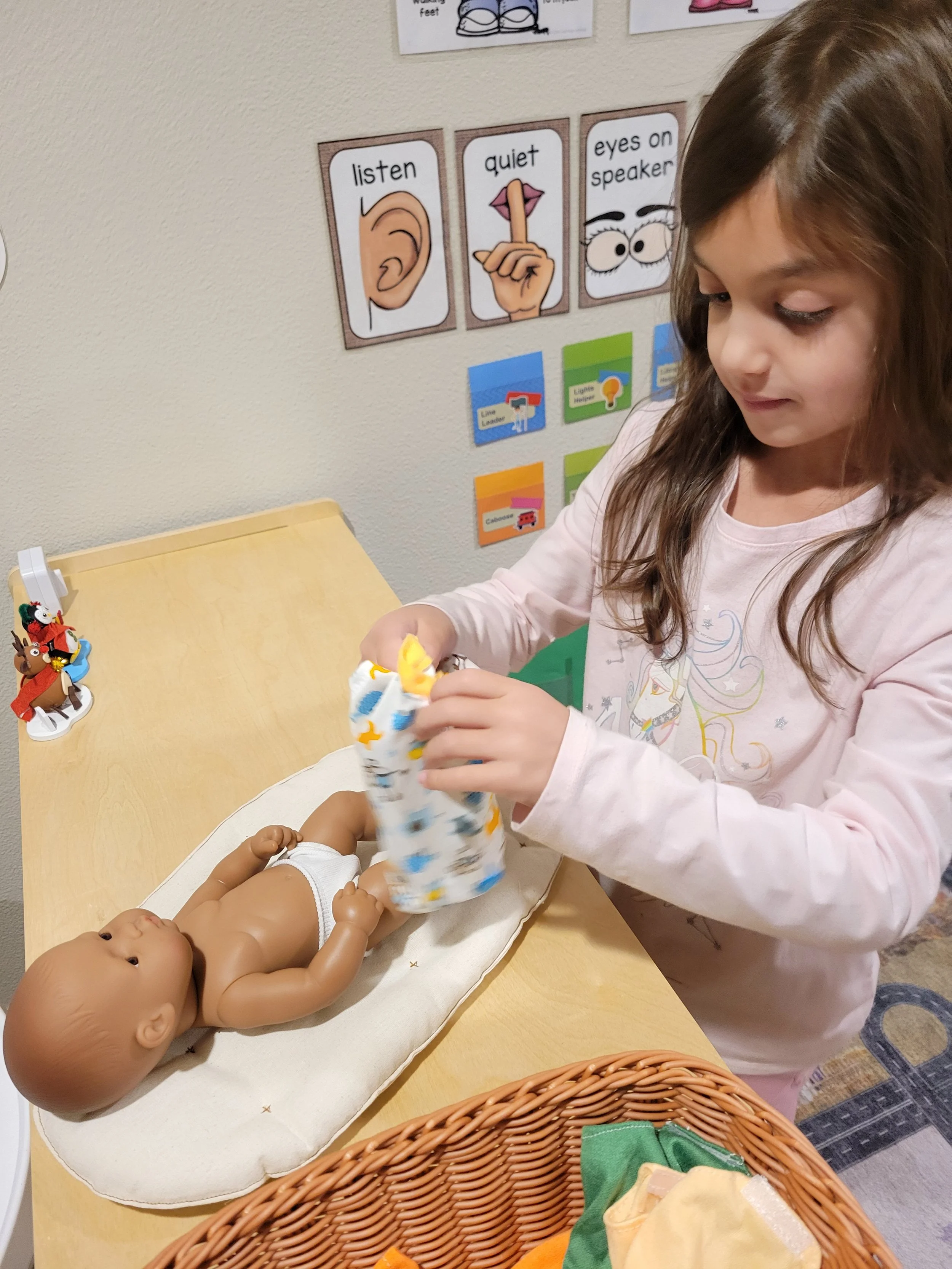 A young girl changing a doll's diaper on a changing pad in a classroom or daycare setting, with educational posters on the wall.