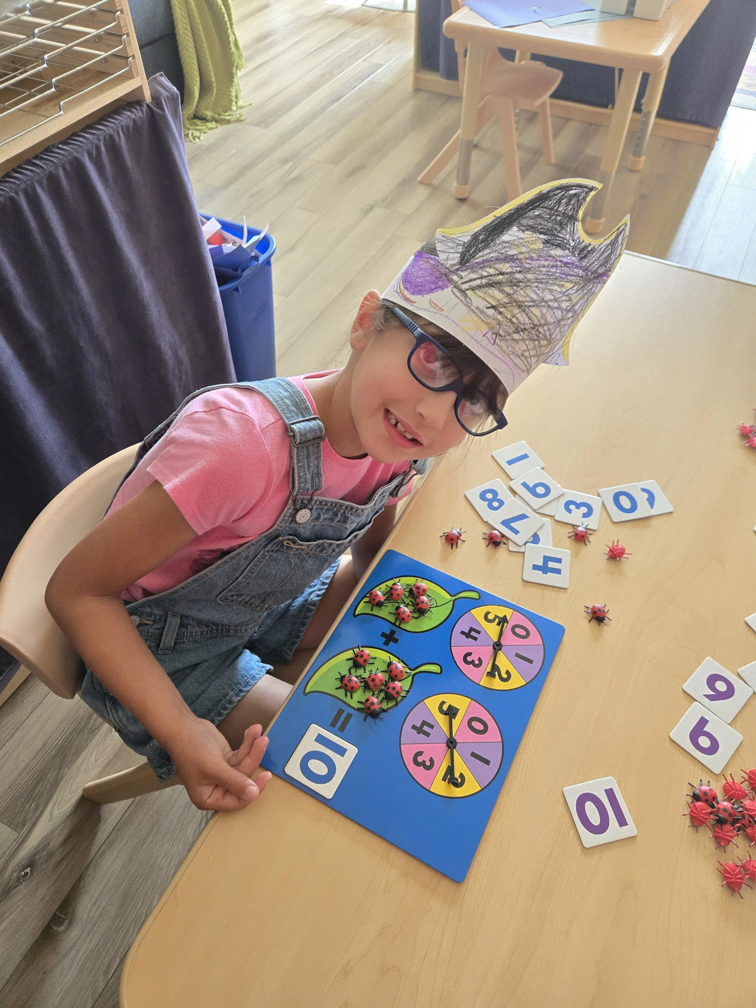 A young girl wearing glasses and a paper crown is sitting at a table with a colorful math game and number tiles. She is smiling and holding one of the number tiles.