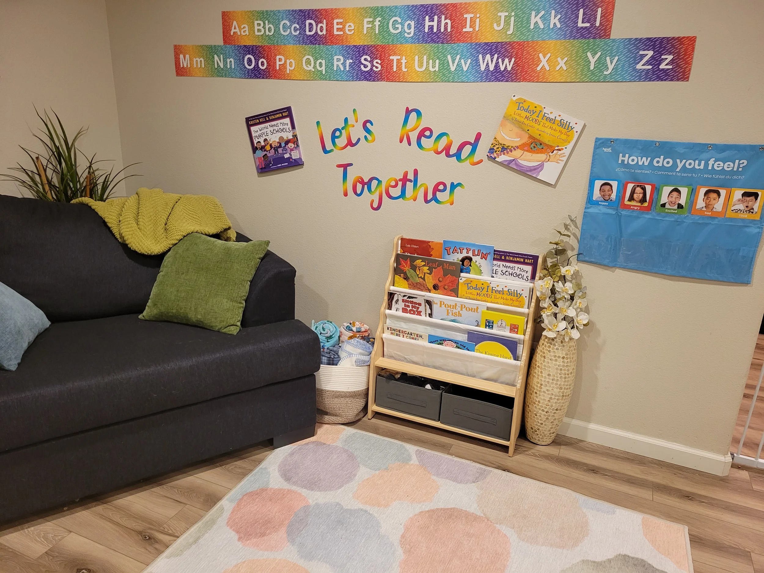 A cozy reading corner in a living room with a dark gray couch, green and blue cushions, a yellow throw blanket, a potted plant, baskets, a wooden book rack with children's books, and colorful wall decorations including alphabet banners, 'Let's Read T