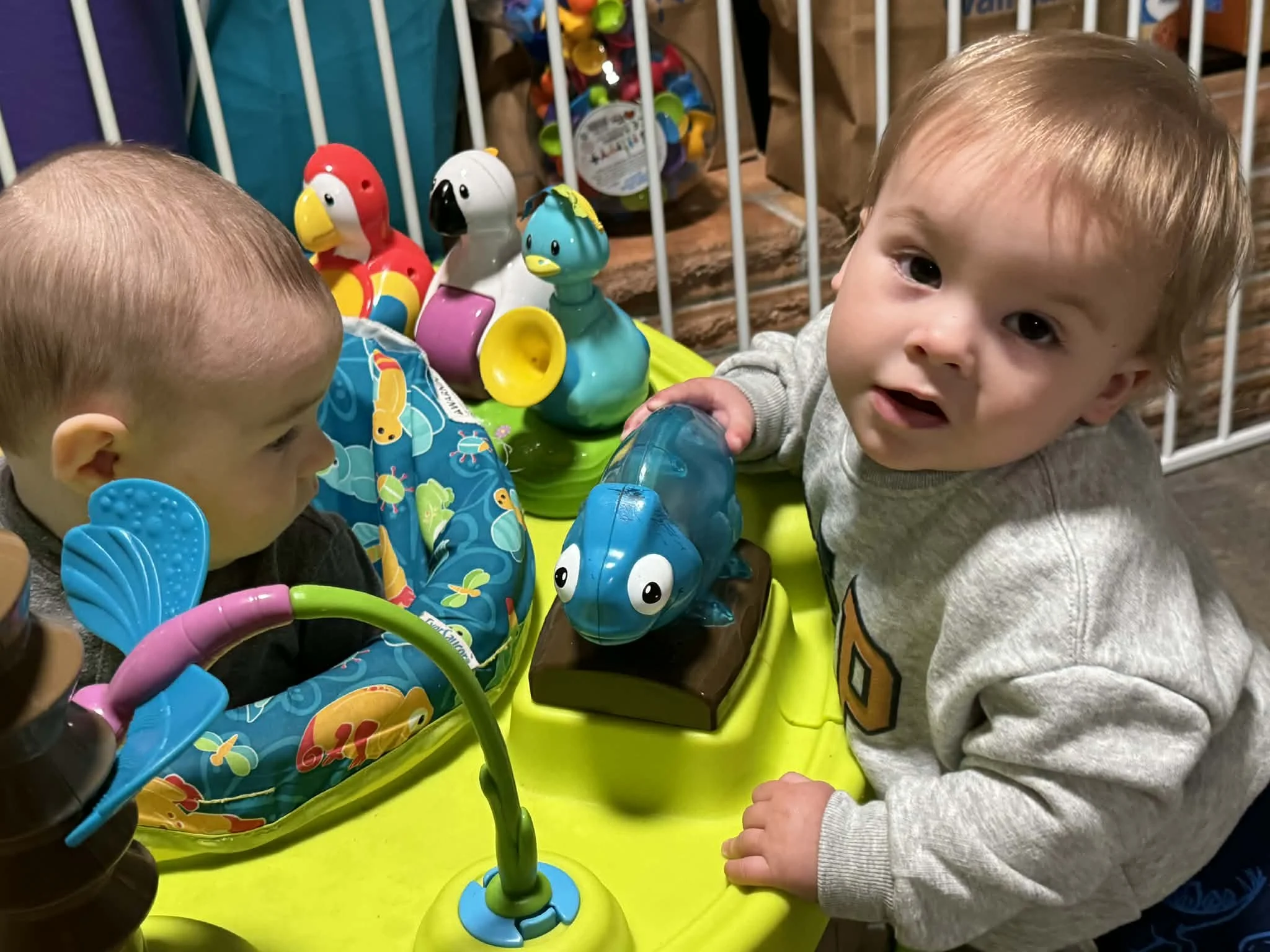 Two young children playing with colorful toys at a bright yellow activity table, with a small baby in a blue and green baby seat.