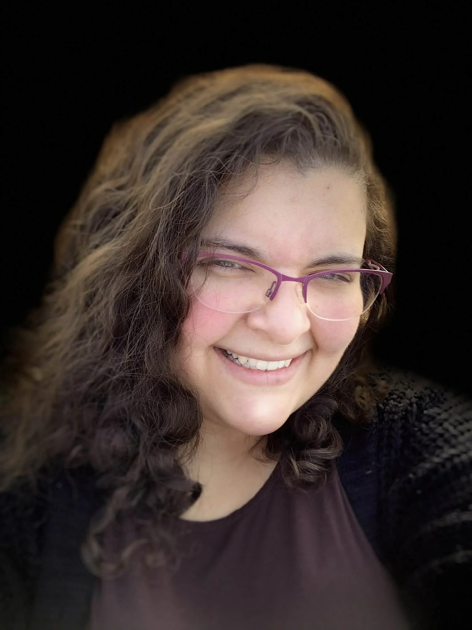 A smiling woman with curly brown hair, wearing purple glasses, a dark top, and a black knit sweater, against a black background.