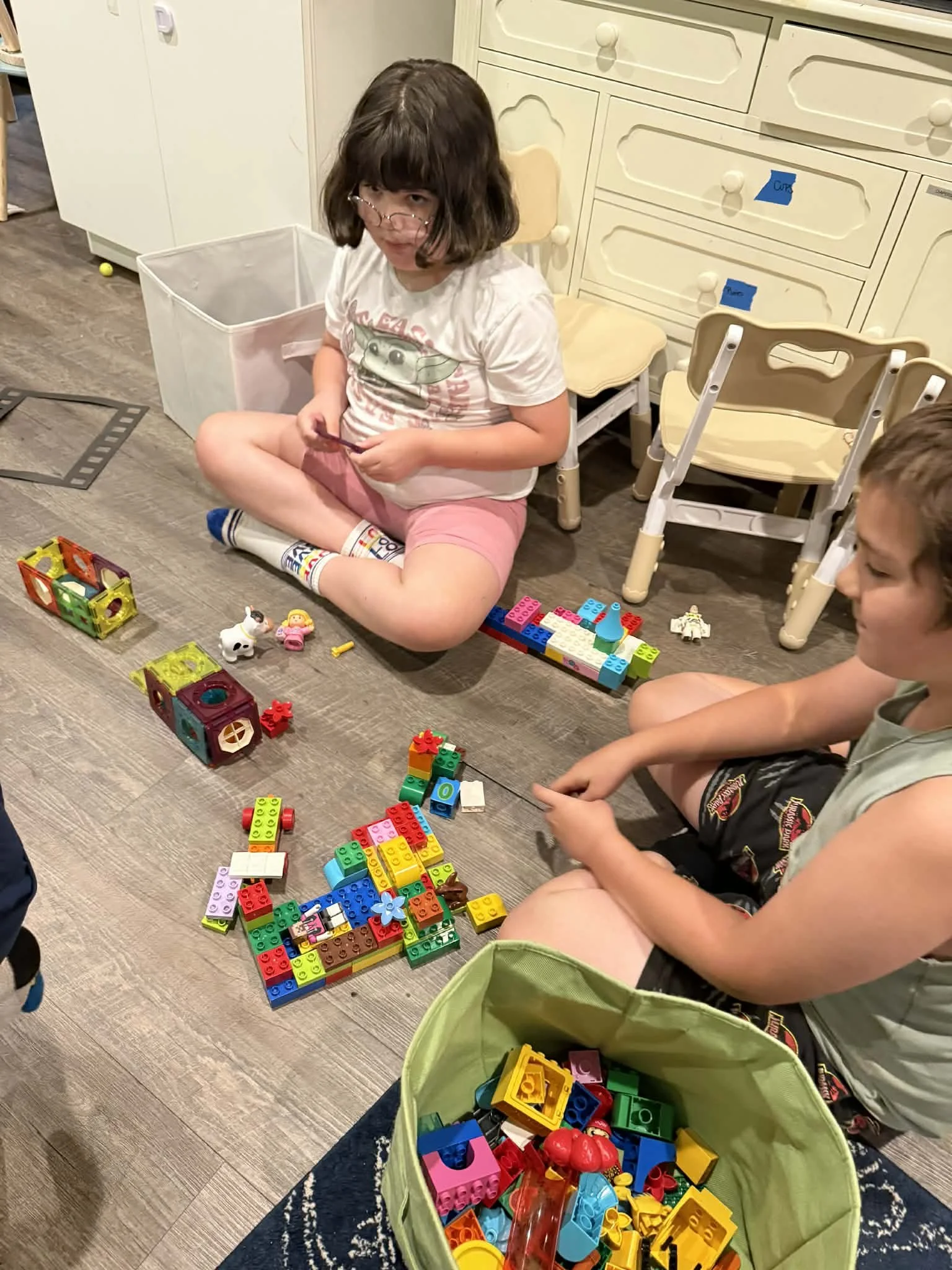 Two children sitting on a wooden floor playing with colorful LEGO blocks, toys, and figures. There are chairs, a storage bin, and kitchen cabinets in the background.