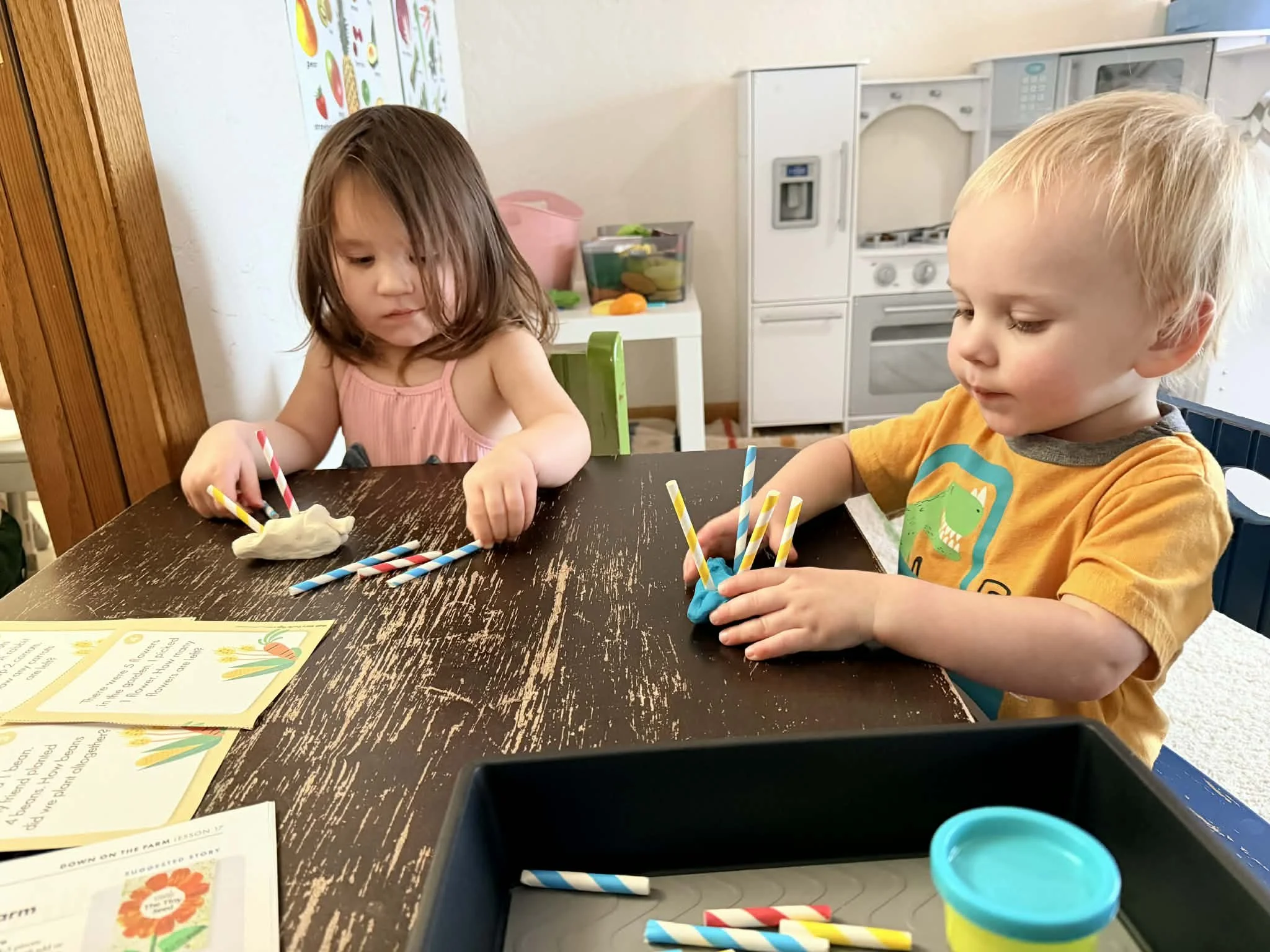 Two young children sitting at a worn wooden table, playing with colorful modeling clay and striped paper straws, in a playroom with children's drawings on the wall and a small kitchen set in the background.