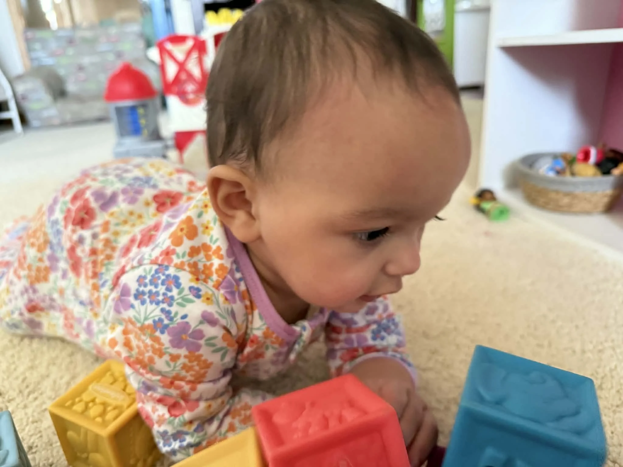 A young child with short hair and wearing a colorful floral long-sleeve shirt is lying on a carpeted floor, playing with plastic blocks. There are baskets and shelves with toys in the background.