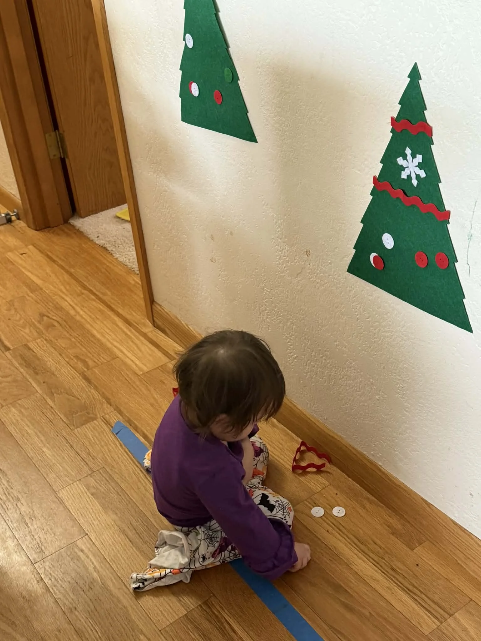 Child sitting on the wooden floor, decorating a wall with Christmas-themed paper cutouts of Christmas trees with ornaments, ribbons, and snowflakes.