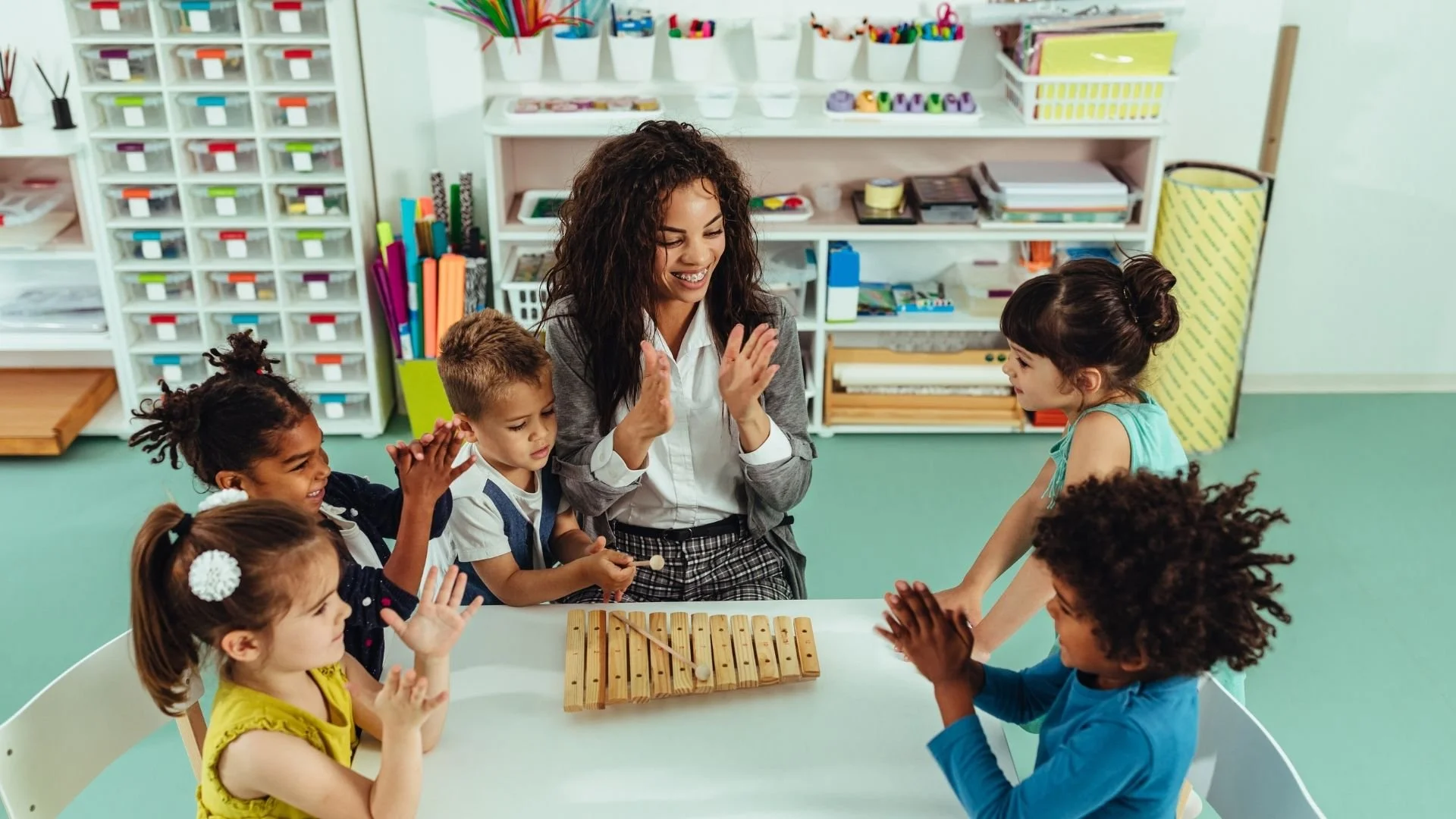 A teacher and six young children sitting around a white table in a classroom, participating in a music activity with a xylophone.