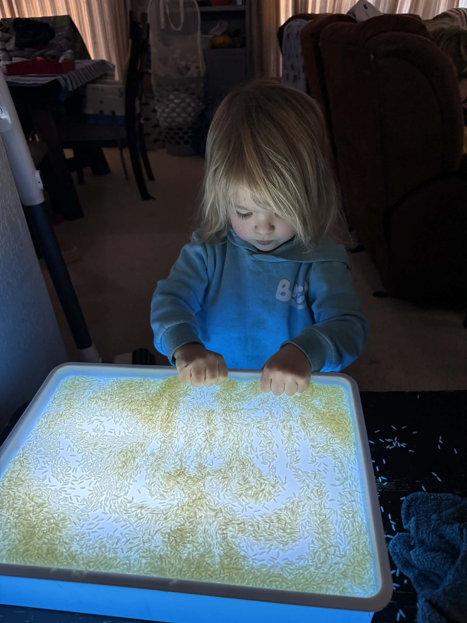 A young child with blonde hair looking down and engaging with an illuminated touch-screen table displaying scattered rice grains or small particles.