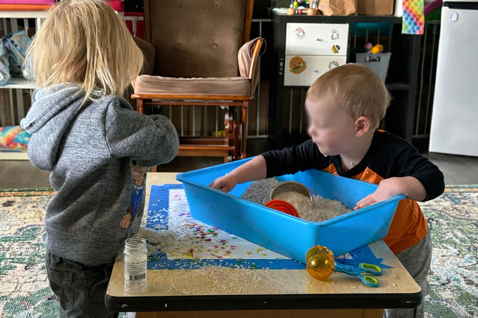 Two children playing with rice and pretend kitchen utensils at a table in a playroom.