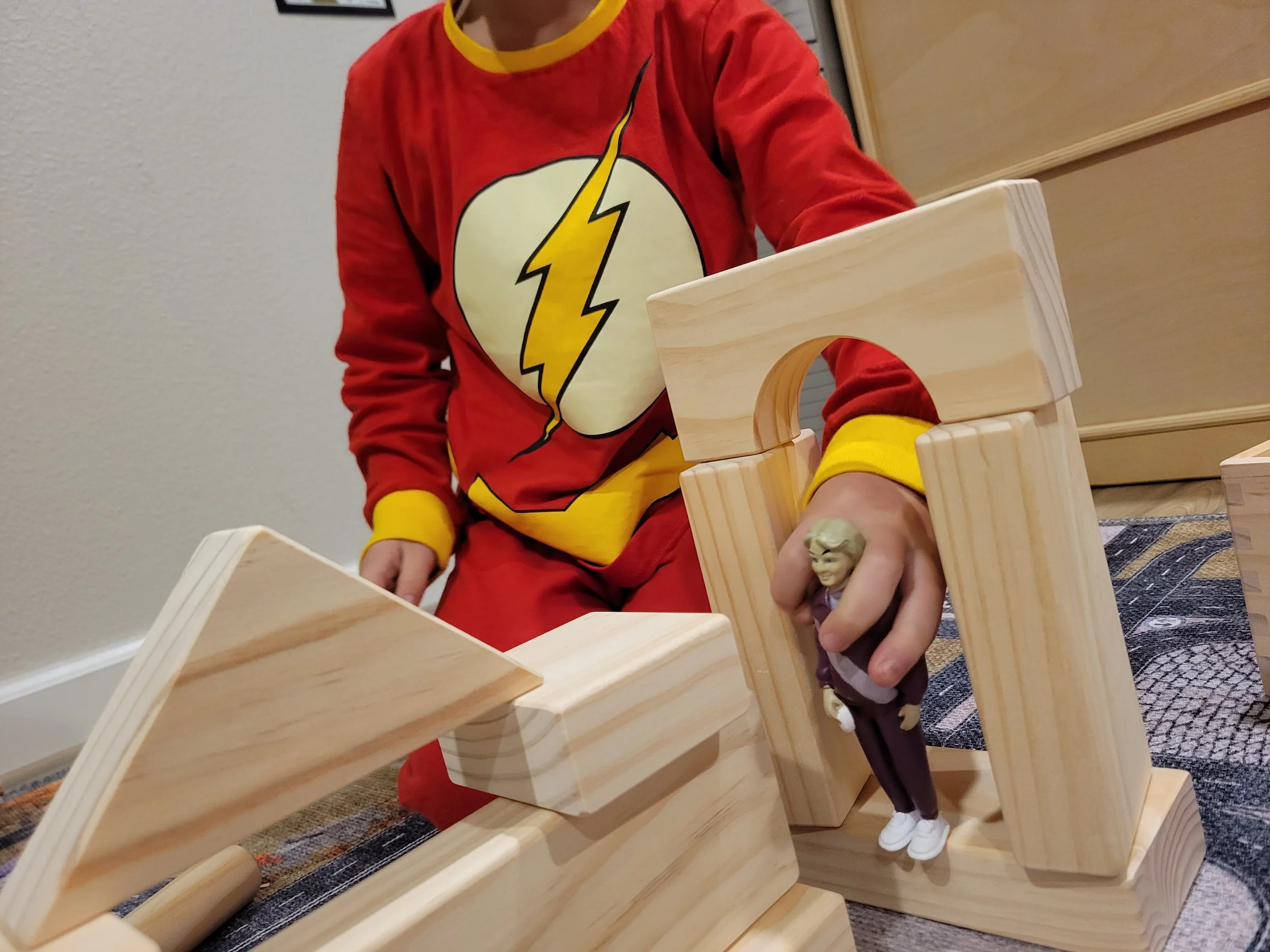 Child in a red and yellow flash-themed pajama set playing with wooden building blocks, holding a small figurine of an elderly person.