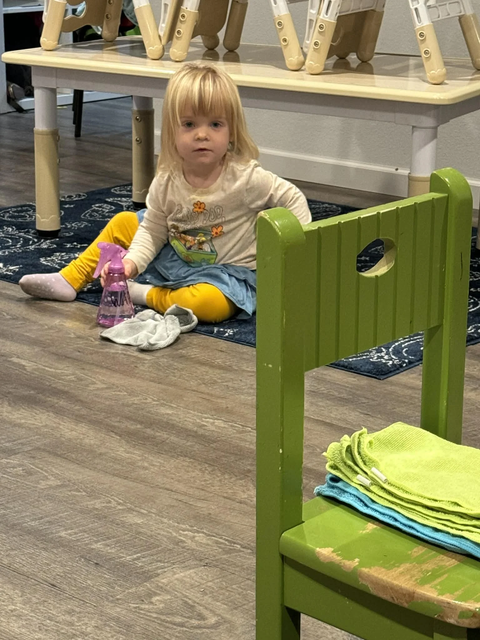 A young girl with blonde hair sitting on the floor, holding a pink spray bottle, near a green chair with stacked yellow and blue towels on it, in a room with wooden flooring and a patterned rug.