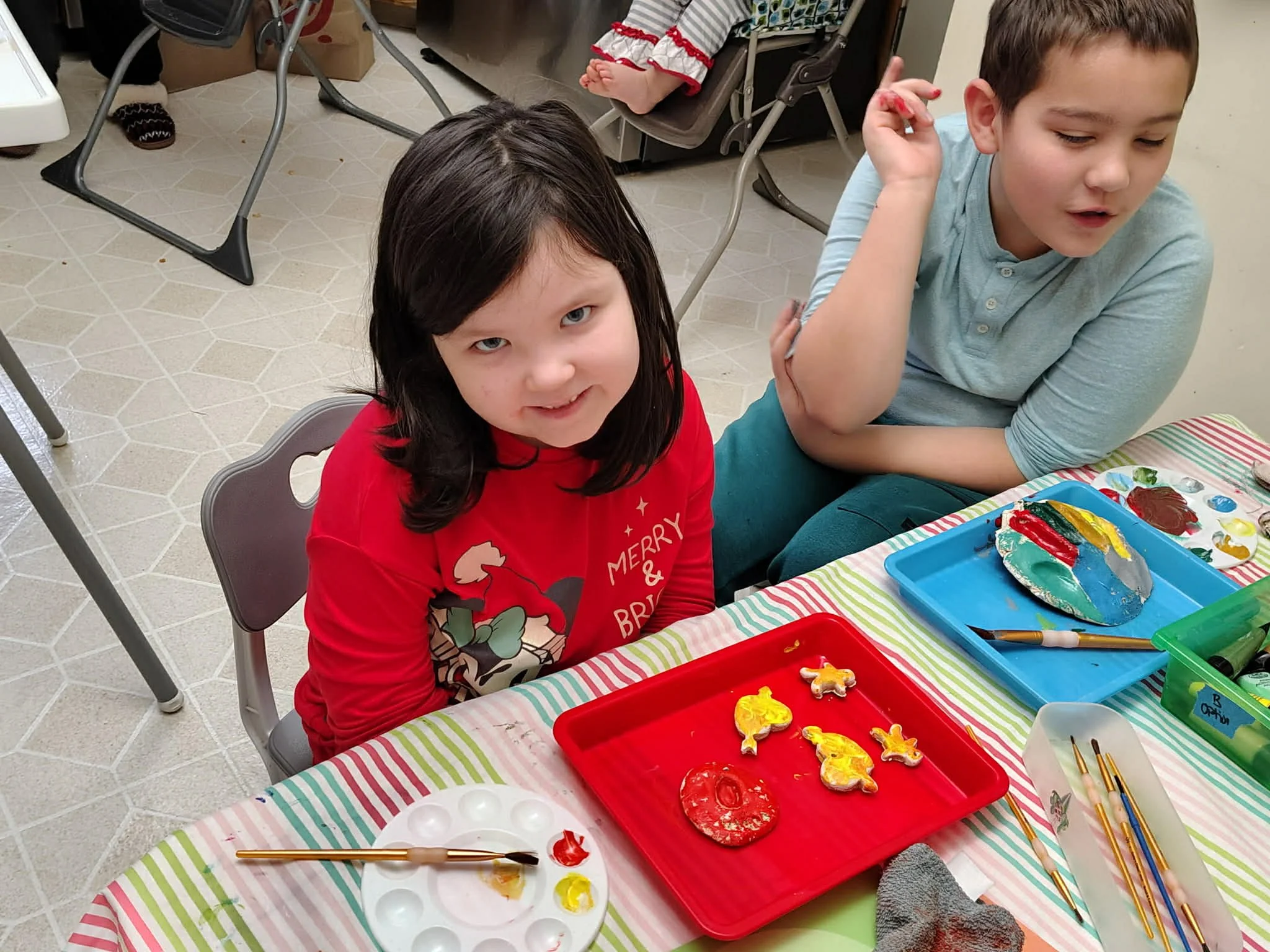 Two children sitting at a table painting holiday ornaments with paint and brushes, surrounded by painting supplies and colorful trays.