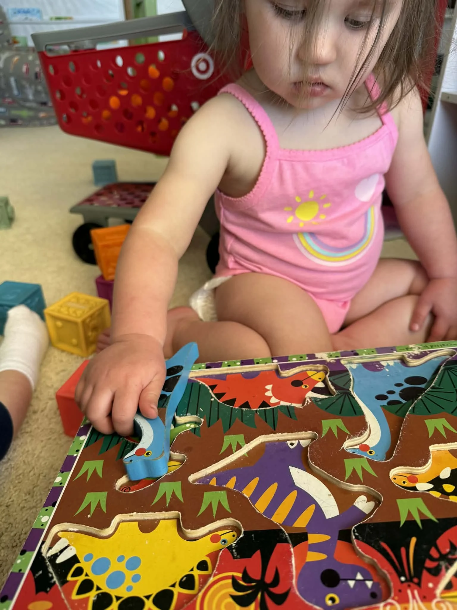 A young girl in a pink tank top with rainbow and sun design, playing with a colorful dinosaur puzzle on the floor of a toy store or play area.