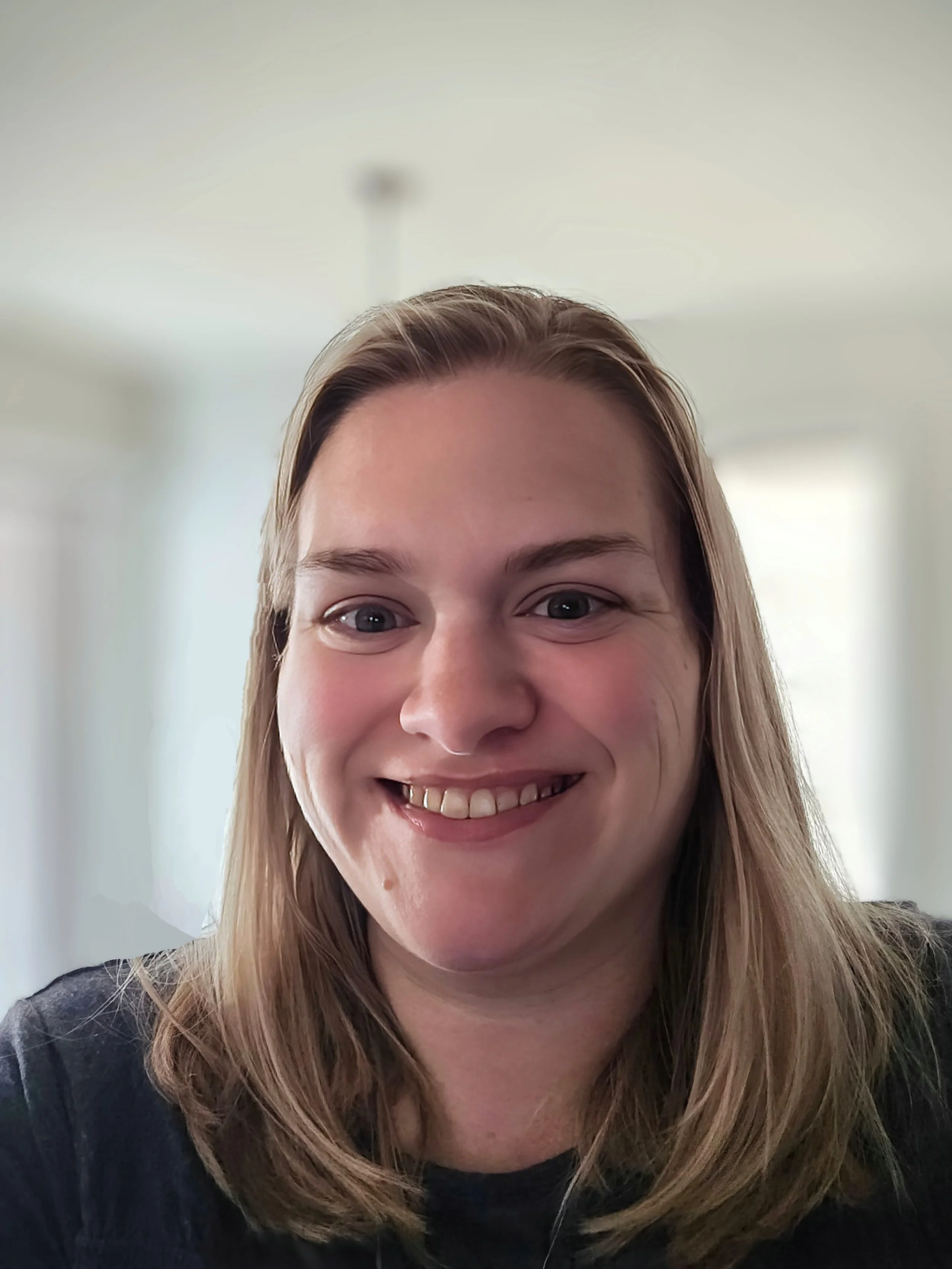 Close-up of a smiling woman with shoulder-length blonde hair, wearing a black top, in a softly lit room with white walls and windows in the background.