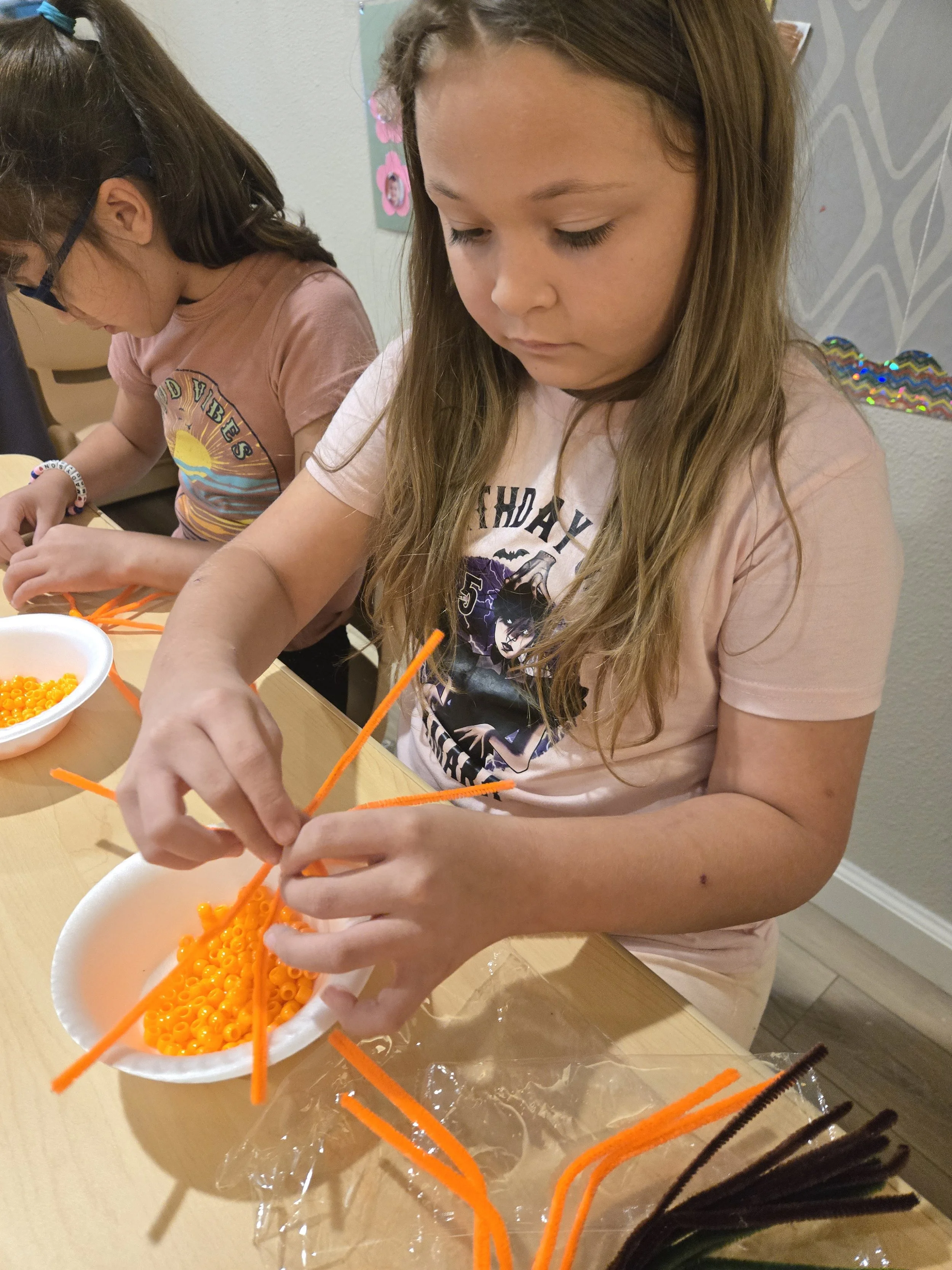 Child making a spider craft with orange and black pipe cleaners and beads at a table.