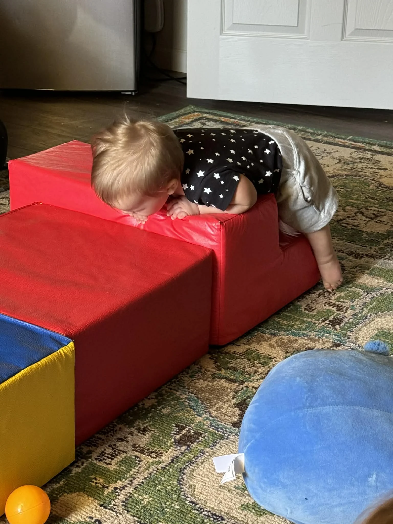 A young child with light brown hair wearing a black shirt with white stars and gray shorts is crawling on a red padded cube and peeking over the edge.