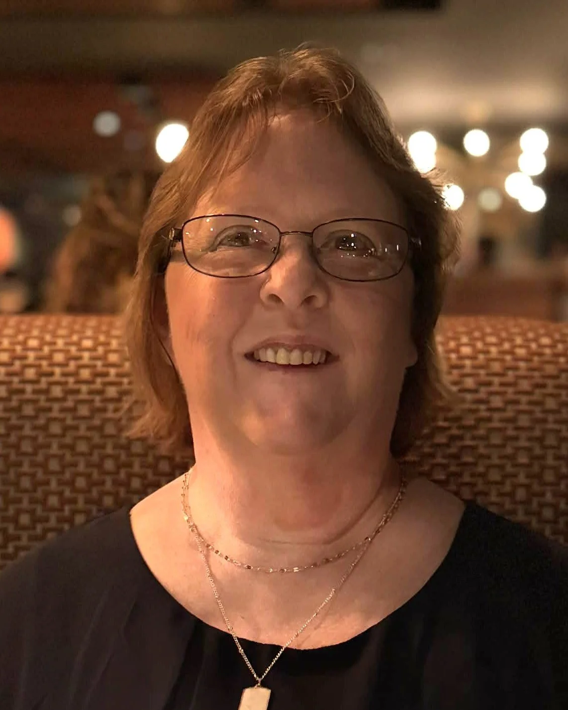 A smiling middle-aged woman with short reddish-brown hair and glasses, wearing two gold necklaces, sitting on a patterned chair in a warmly lit restaurant or cafe.