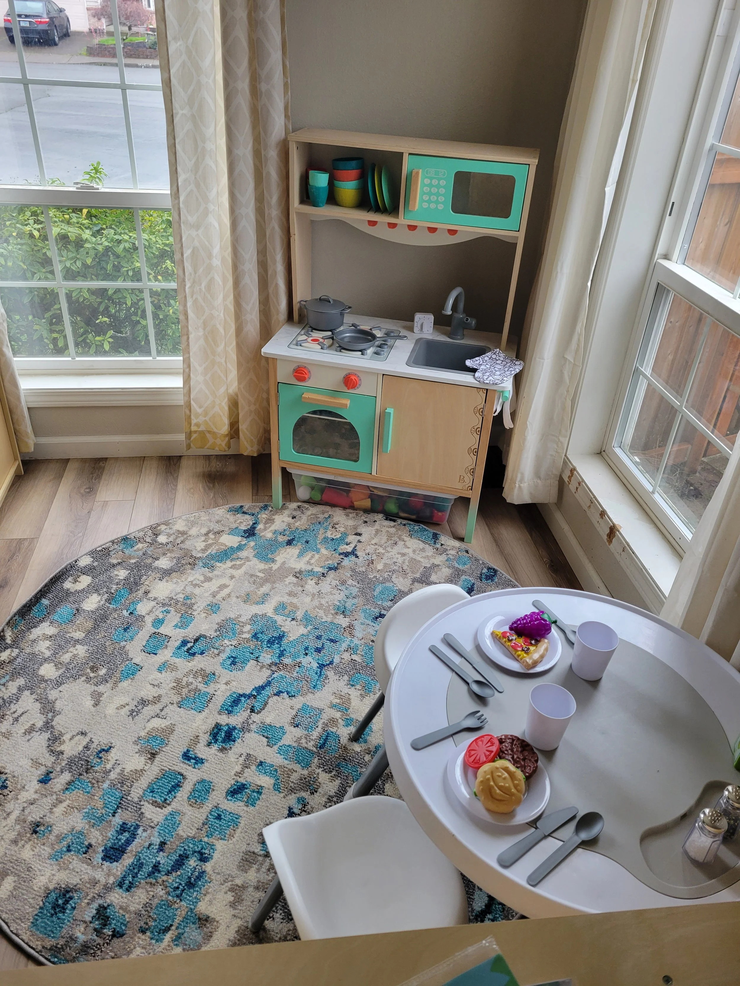 A children's play kitchen with toy appliances and dishes, placed near large windows with beige and white curtains, and a round table set with toy food, cups, and utensils in a bright, cozy room.