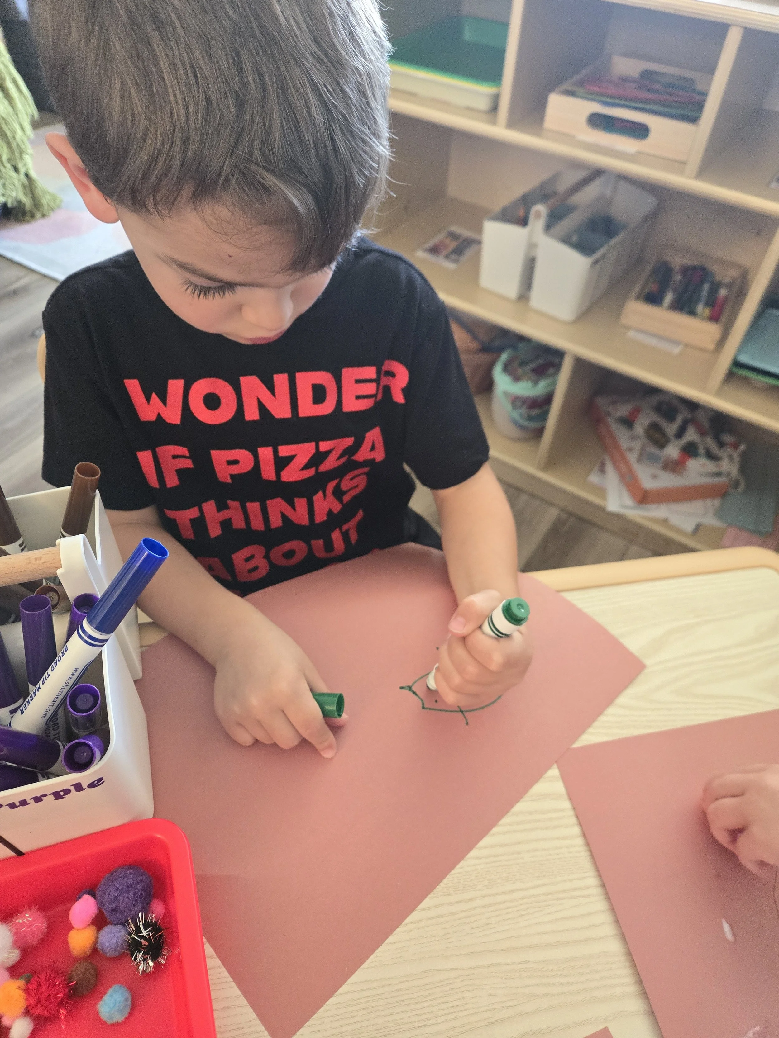 A young boy with brown hair drawing on pink paper with a green marker inside a classroom. There are craft supplies including markers and pom-poms on the table.