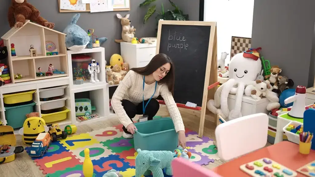 A woman in a cream sweater is sitting on the colorful alphabet rug in a playroom, handling toys from a teal storage bin. The room is filled with stuffed animals, toys, and shelves with organized bins.