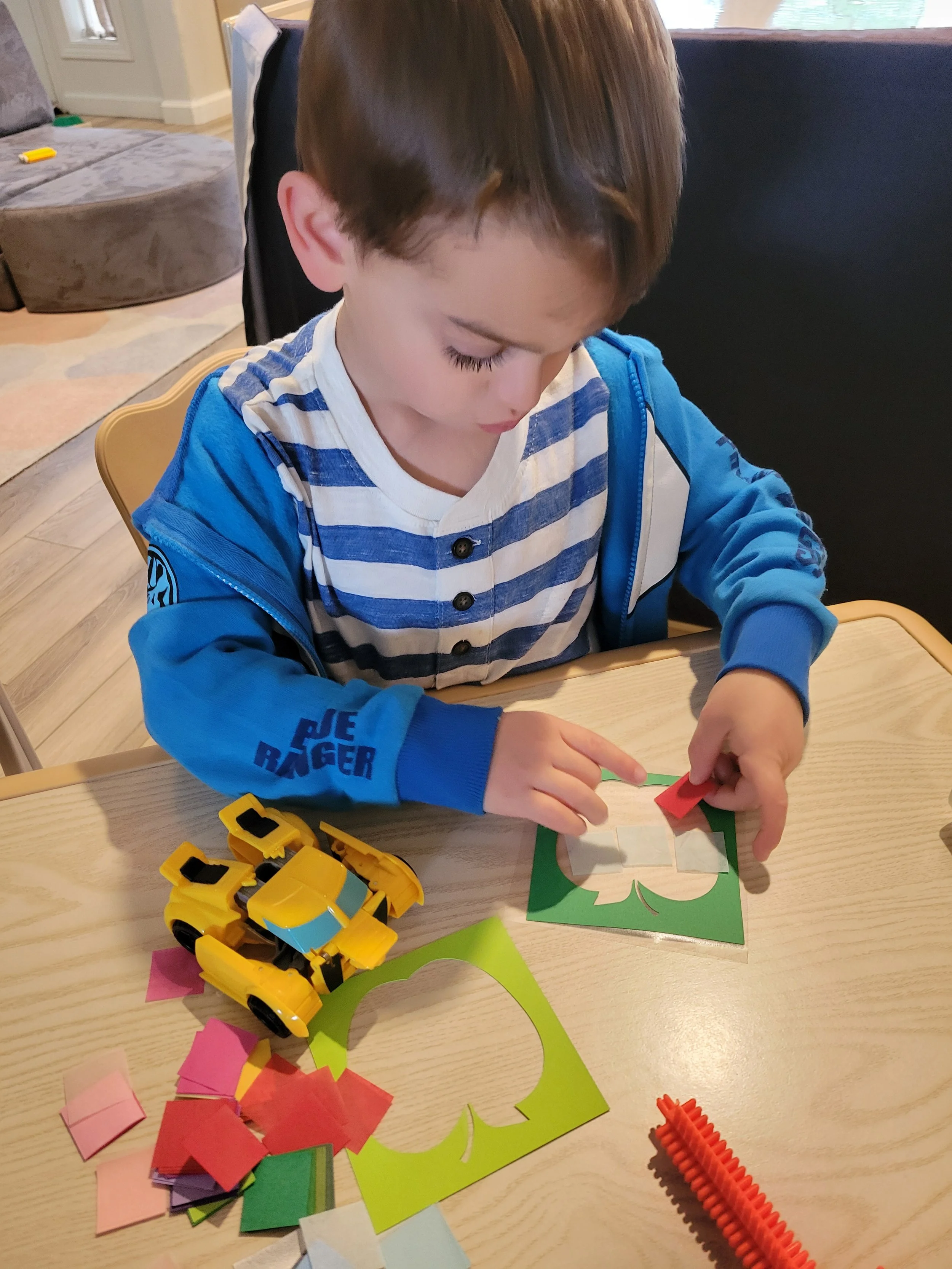 A young boy with brown hair, wearing a blue and white striped shirt and a blue jacket, sits at a wooden table, playing with colorful paper and a yellow toy car. He is folding a piece of paper, surrounded by various small paper squares and a red inter