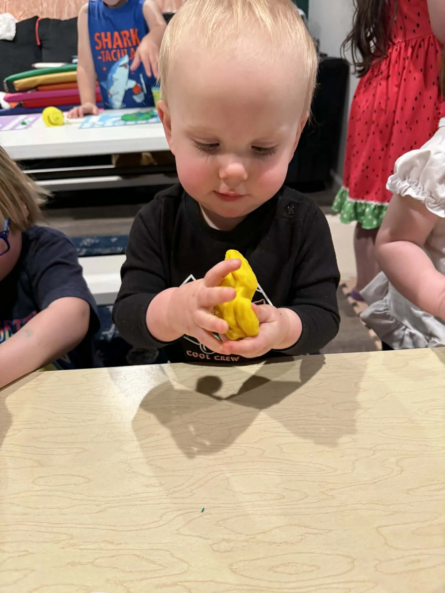 A young boy with blond hair is sitting at a wooden table, playing with yellow playdough. Other children are partially visible around him, with a background of toys and colorful items.