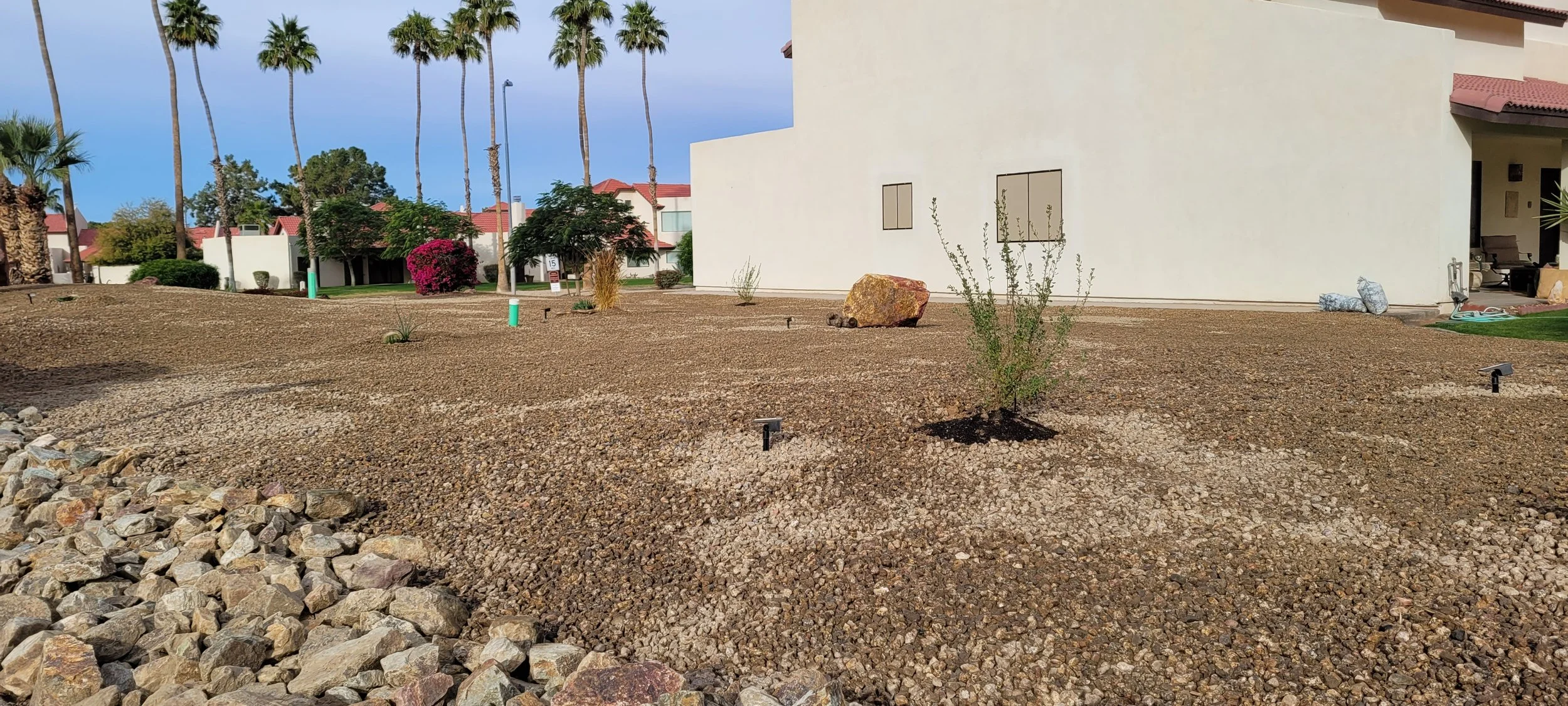 Large rocks called rip rap surrounding smaller decorative gravel rocks in palomino gold coloring with boulders, plants, and solar lighting fixtures in a nice neighborhood of condos.