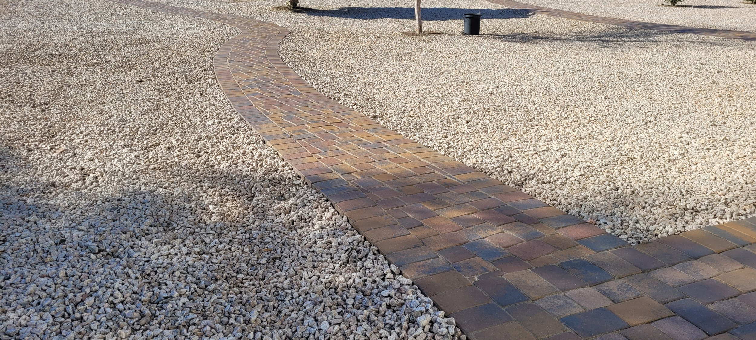 A concrete paver path sealed with concrete sealer in satin finish. The colors are vibrant from Phoenix Pavers, Navajo Blend. It has hues of yellow, orange, reds, grays and tans. There is much decorative white gravel around the path.