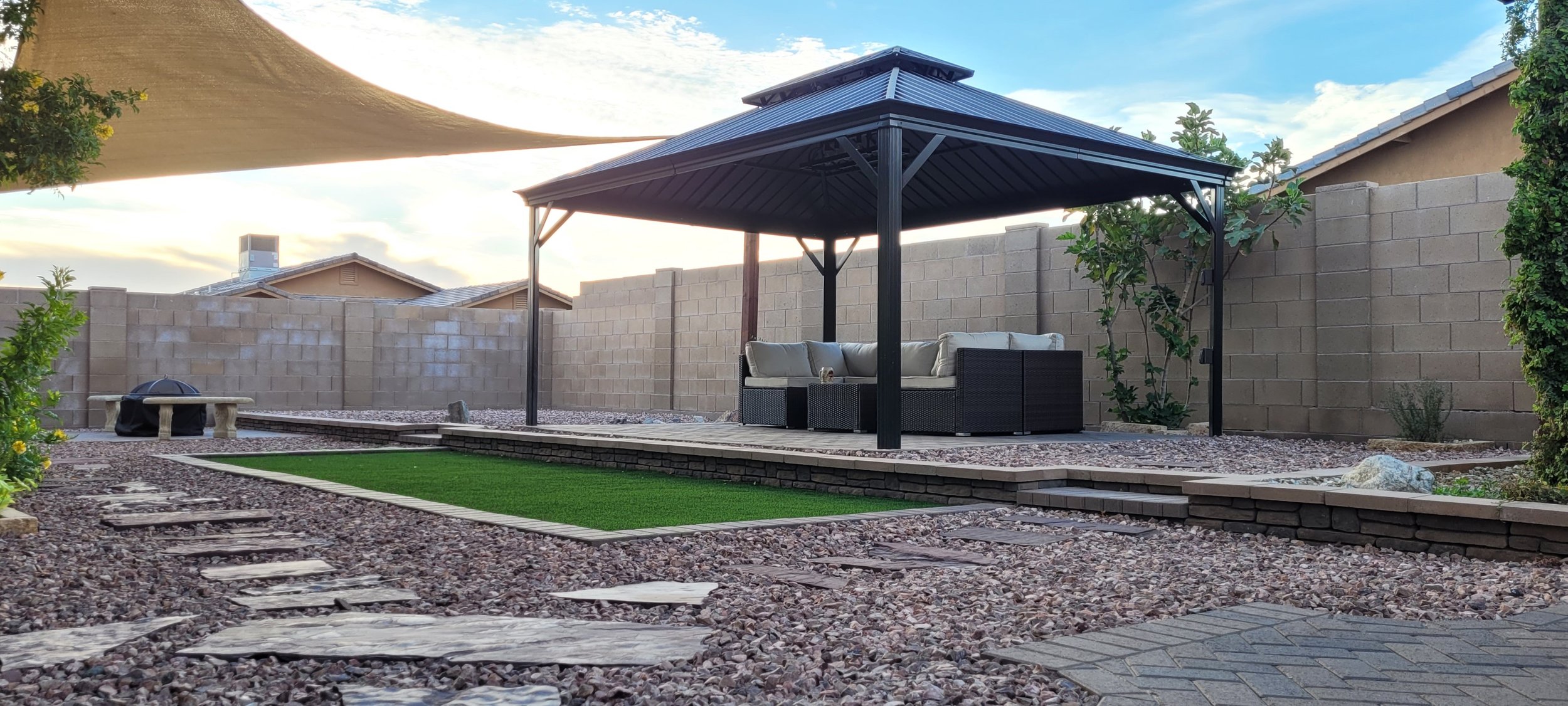 A black gazebo with outdoor furniture. The furniture is dark brown rattan with ivory cushions and a small table in the middle. There is artificial turf with paver edging and a path of flagstone. There is a retaining wall and a shade sail. Cohesive.