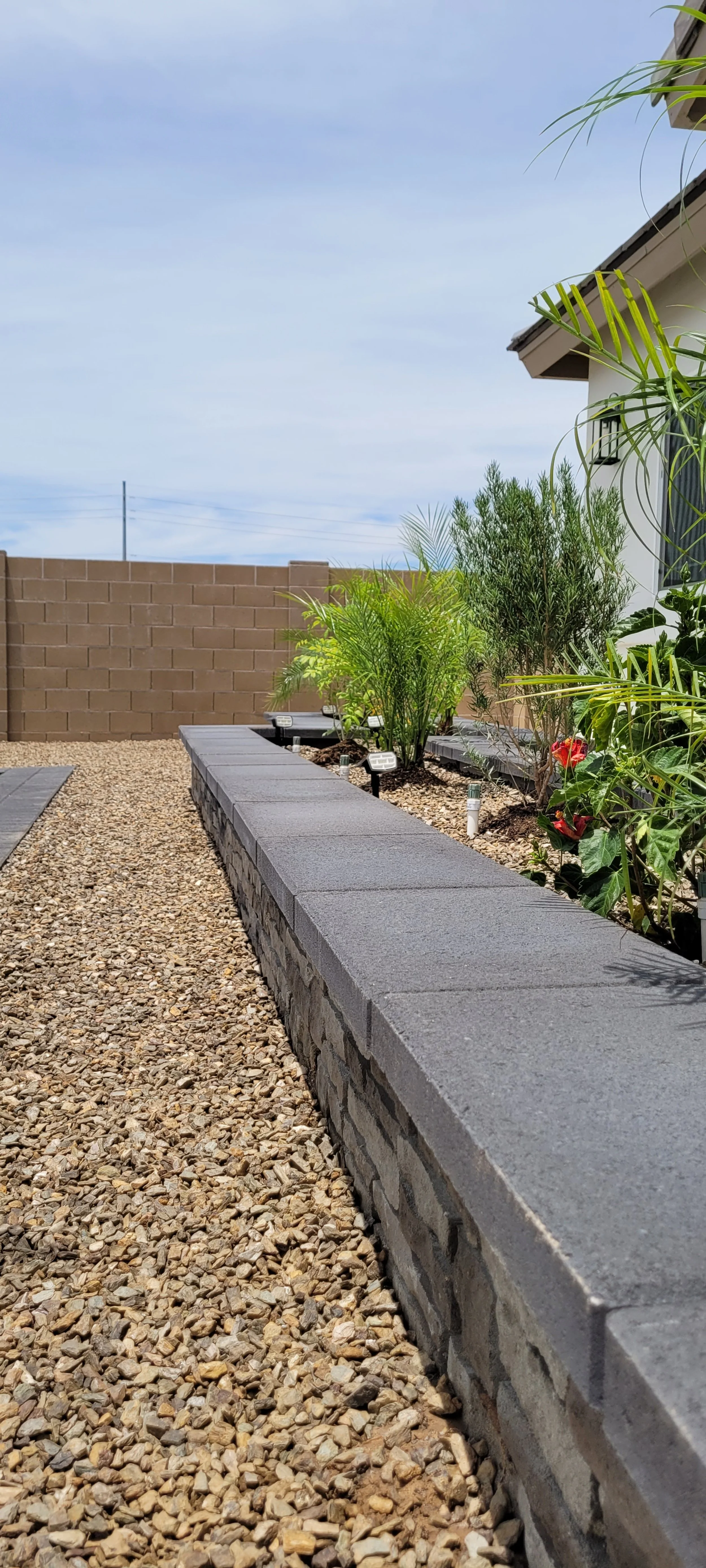 Graphite and dark gray colored retaining wall planter with plants inside. The plants have water bubblers, solar lighting fixtures to light them up at night, and decorative rock in the planter. The plants have soil mulch.