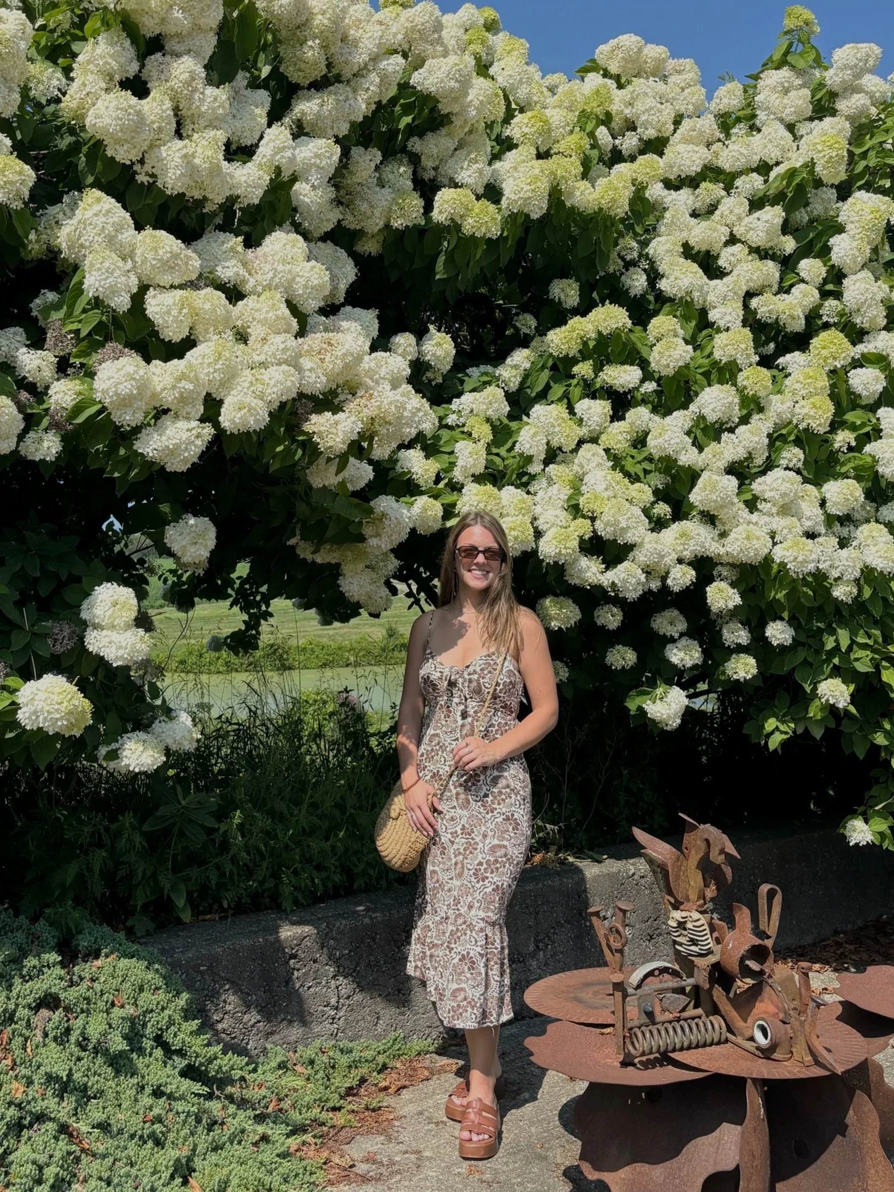 A woman with sunglasses and a floral dress stands in front of a large bush with white flowers. She is holding a woven bag and smiling. There is garden art made of rusted metal nearby, and lush greenery in the background under a blue sky.