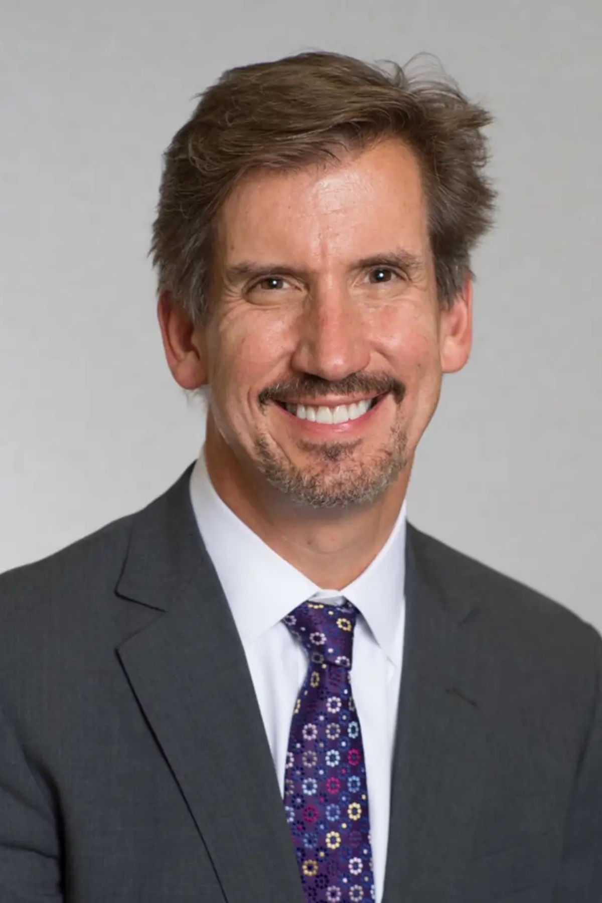 Professional portrait of Dr. Peter A. Zahos, board-certified neurosurgeon, wearing a dark suit, white shirt, and patterned purple tie.