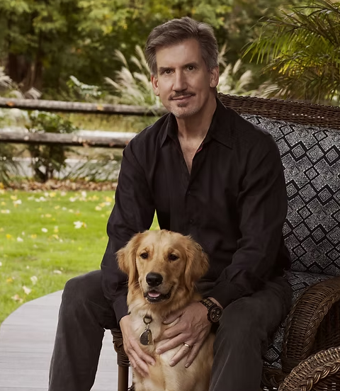 Headshot of Dr. Peter A. Zahos, neurosurgeon, dressed in black dress clothes outdoors on a wicker chair with a golden retriever dog, surrounded by greenery and trees.