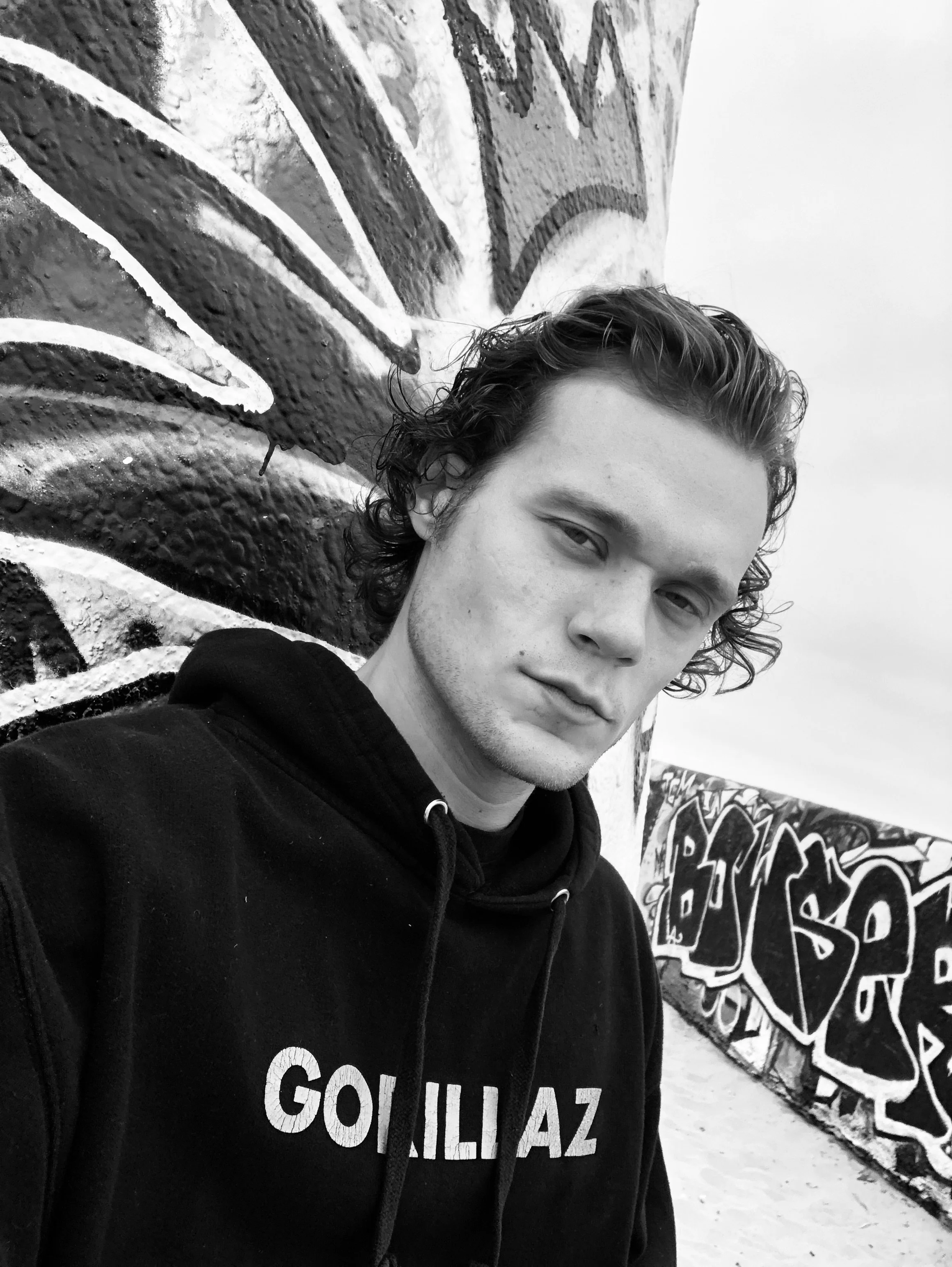 Black and white photo of a young man with curly hair taking a selfie in front of graffiti-covered walls.