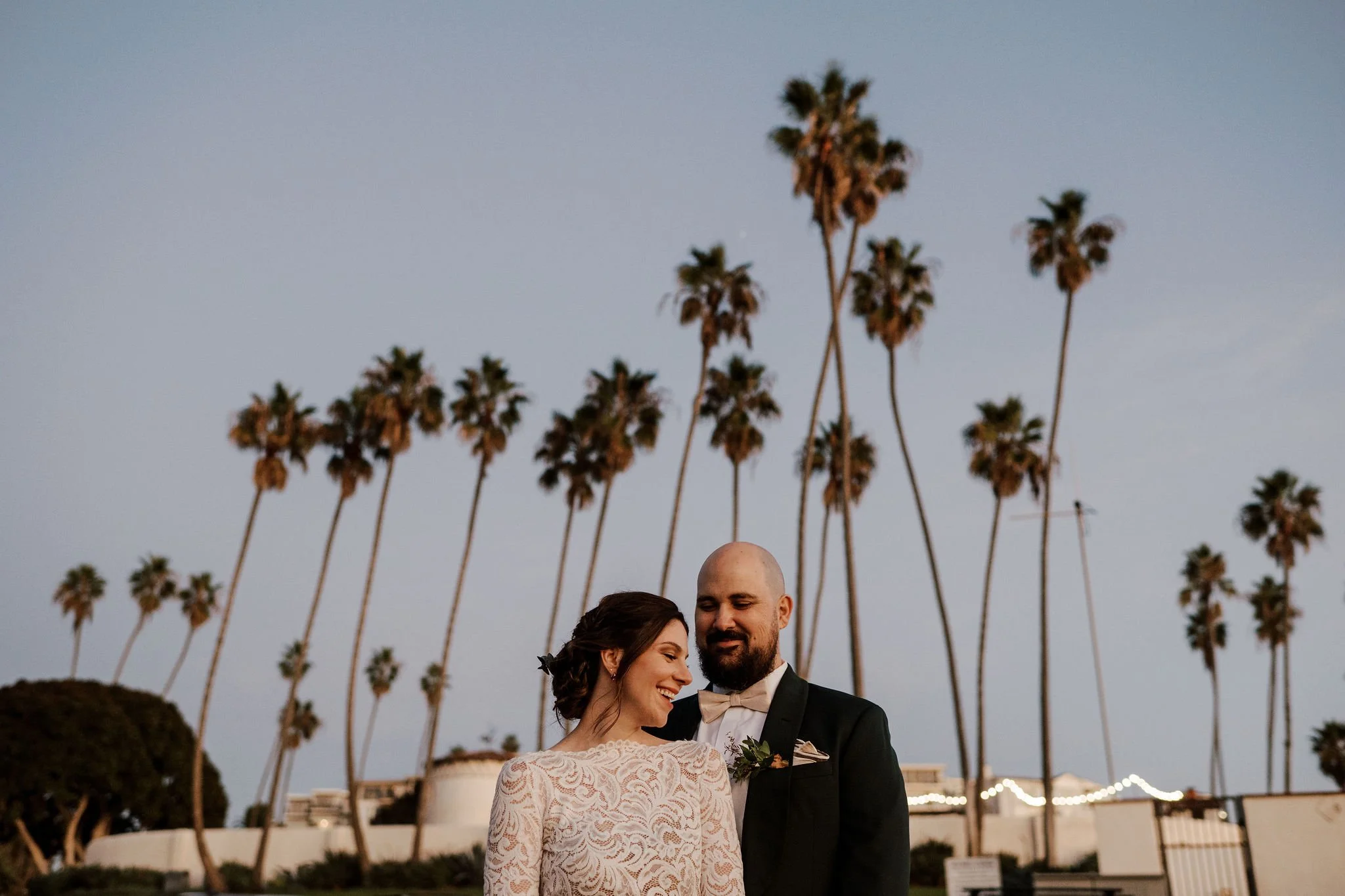 A bride and groom pair smiling and standing close together outdoors during sunset, with tall palm trees in the background.